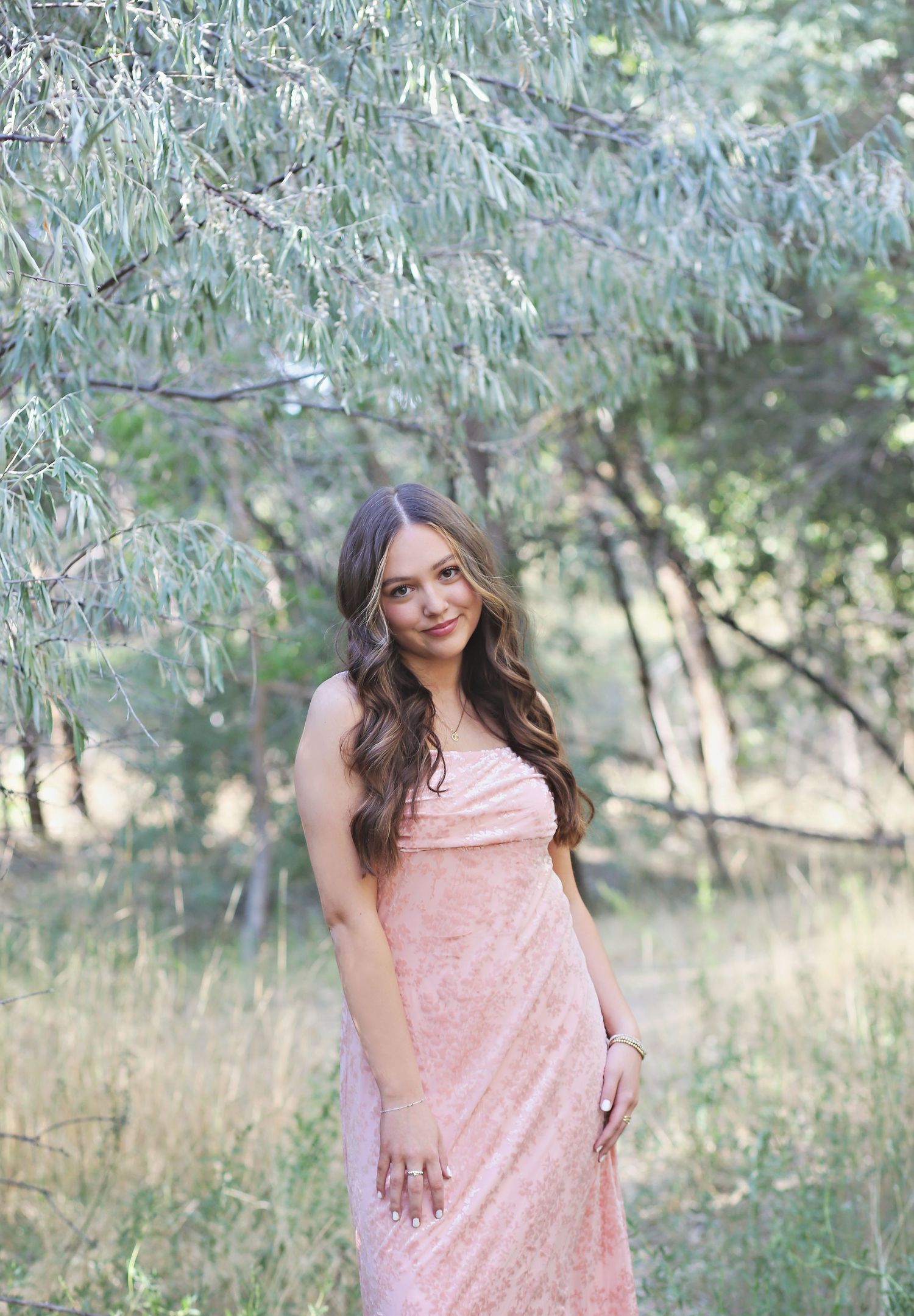 Person in pink dress poses outdoors among olive trees in natural lighting.