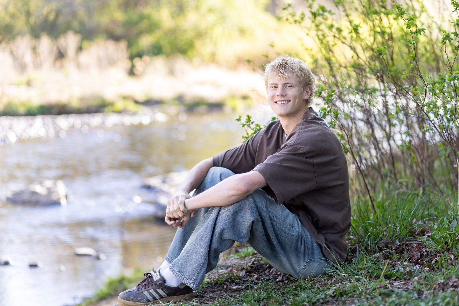 Smiling teenage boy with blonde hair sitting outdoors by a creek, wearing jeans and a brown shirt.