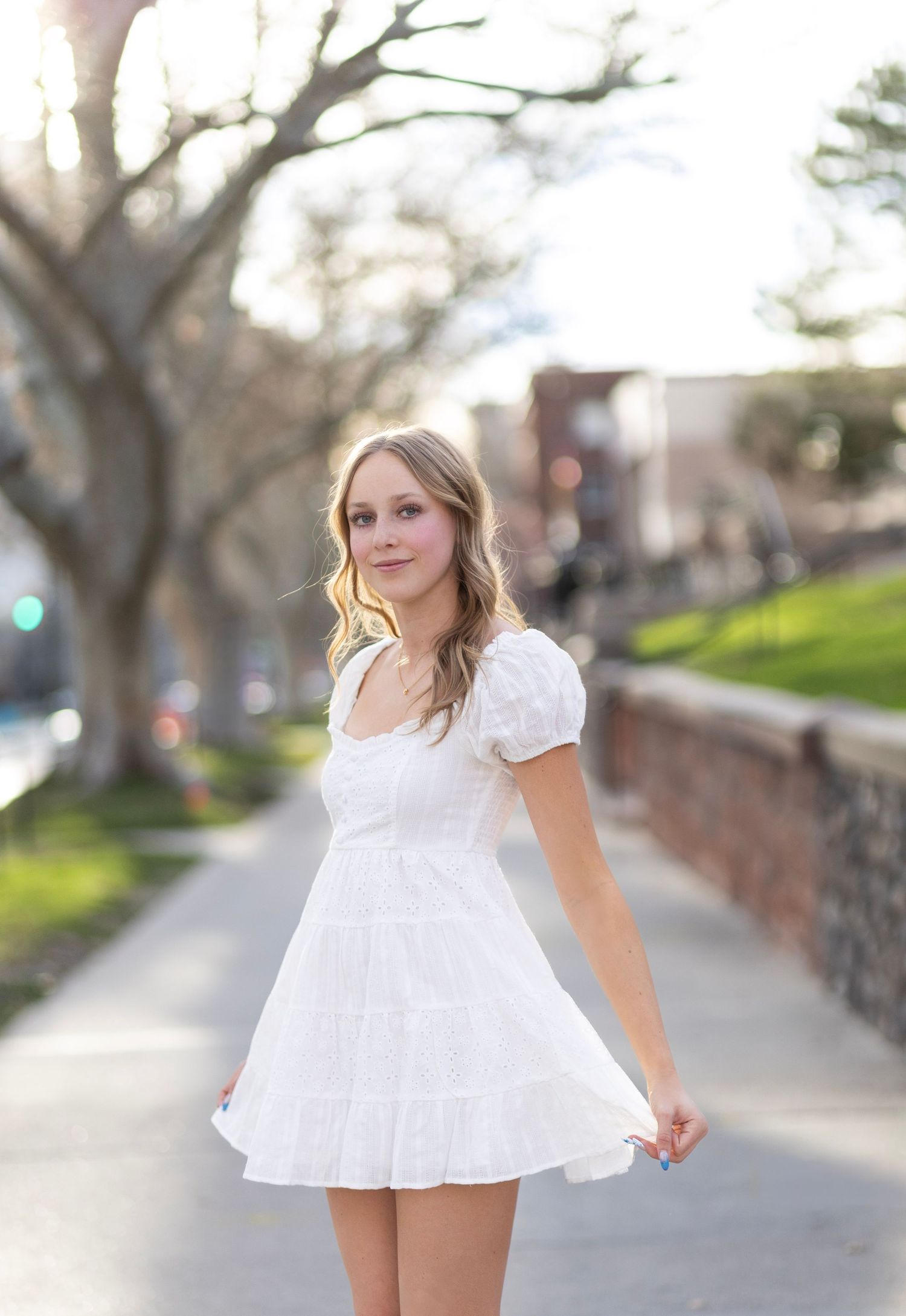 Young woman in white mini dress posing on a sunny sidewalk surrounded by trees and brick buildings.