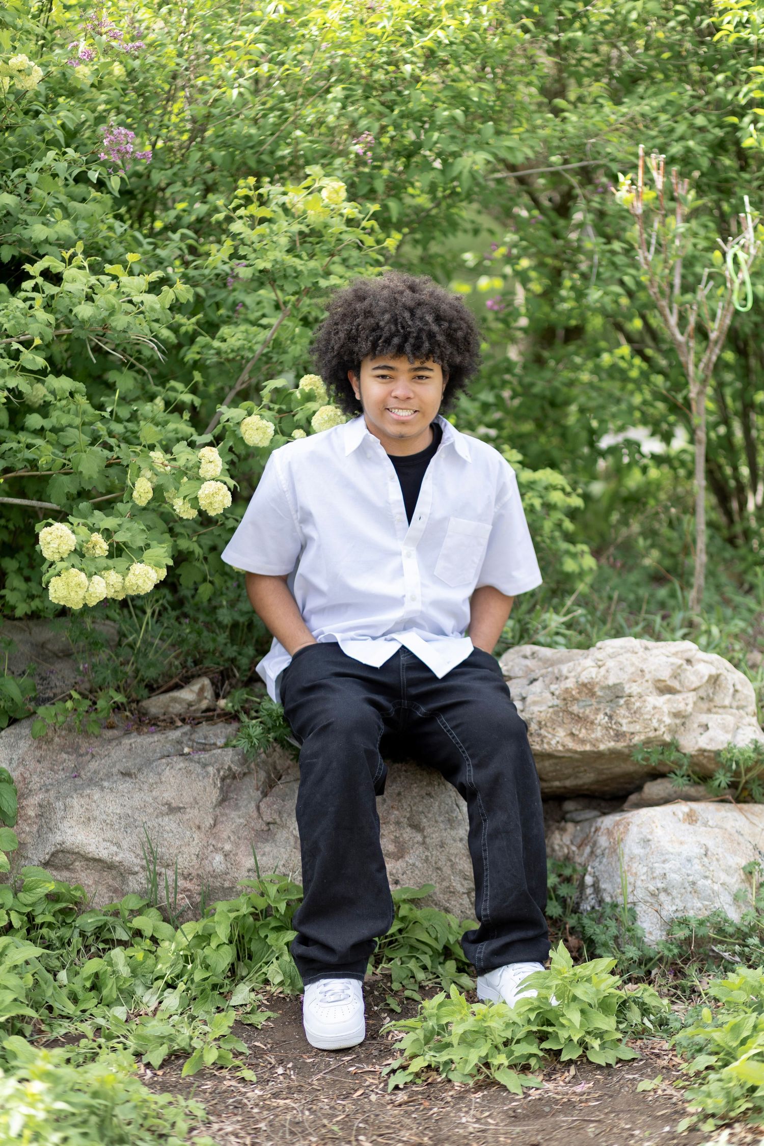 Young teen boy with curly hair sits on rocks in a lush garden, wearing a white shirt and black pants.