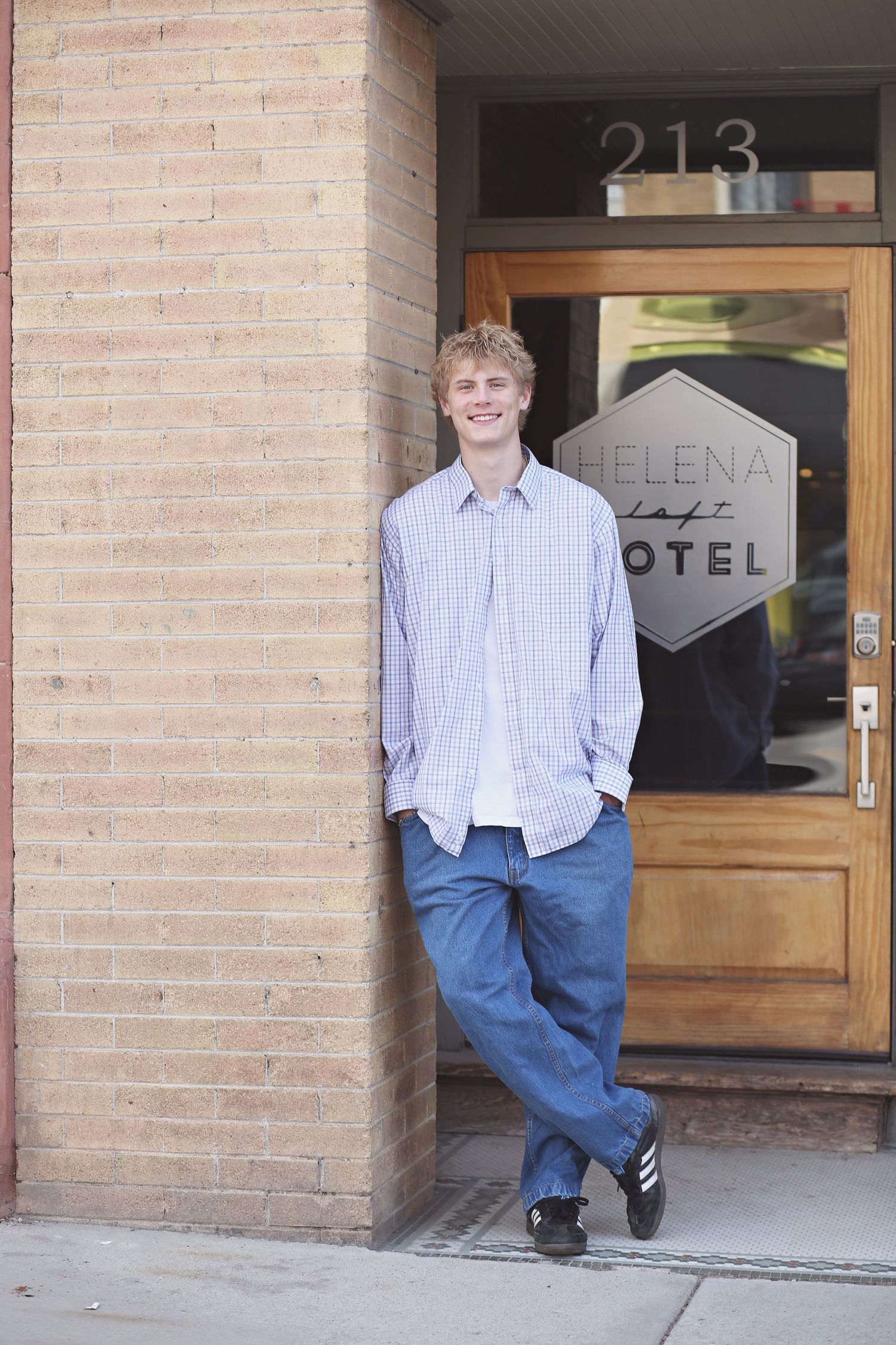 A person in a light blue shirt and jeans stands casually against a brick wall outside a building entrance.