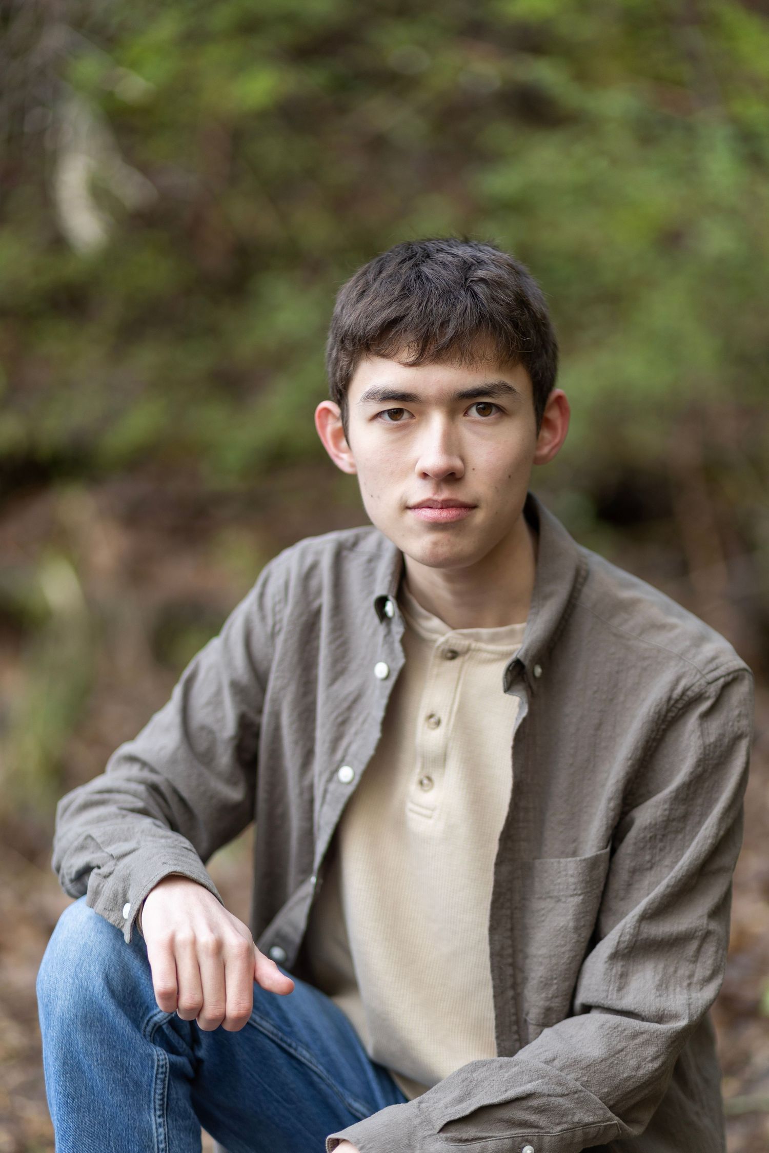 Young man in gray shirt and jeans sitting outdoors with green foliage background, casual portrait photo.