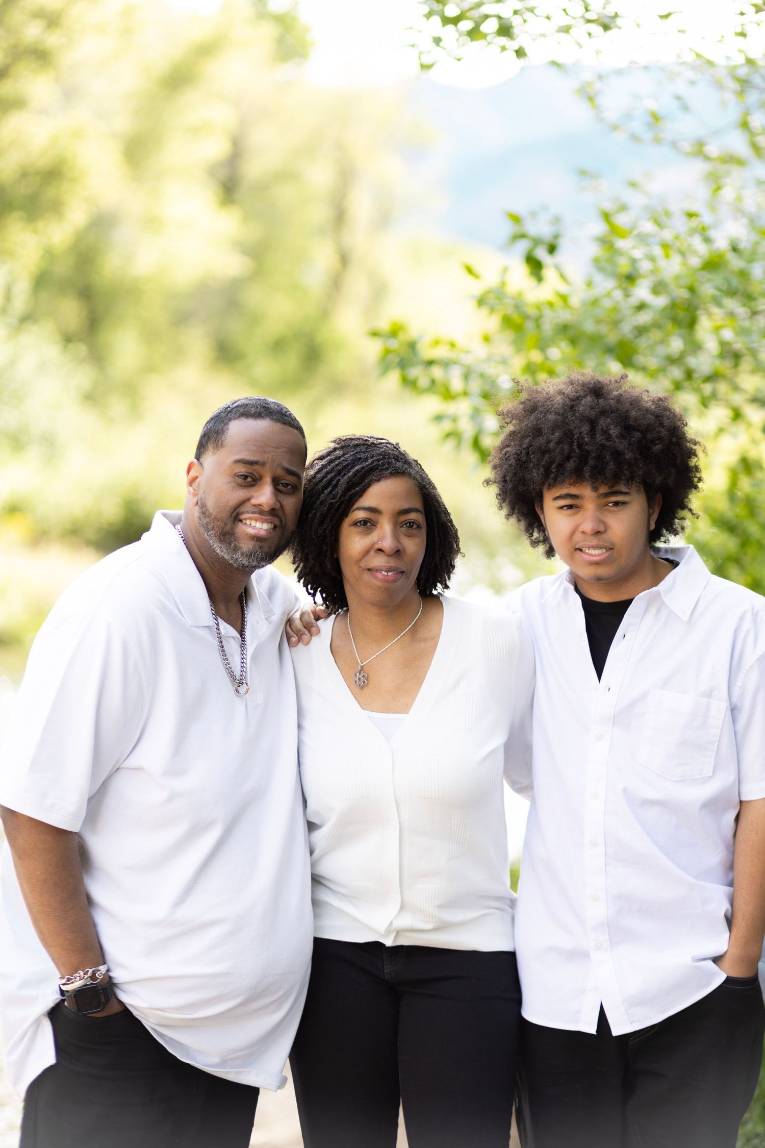 Happy African American family of three posing outdoors in white shirts, smiling together surrounded by green trees.