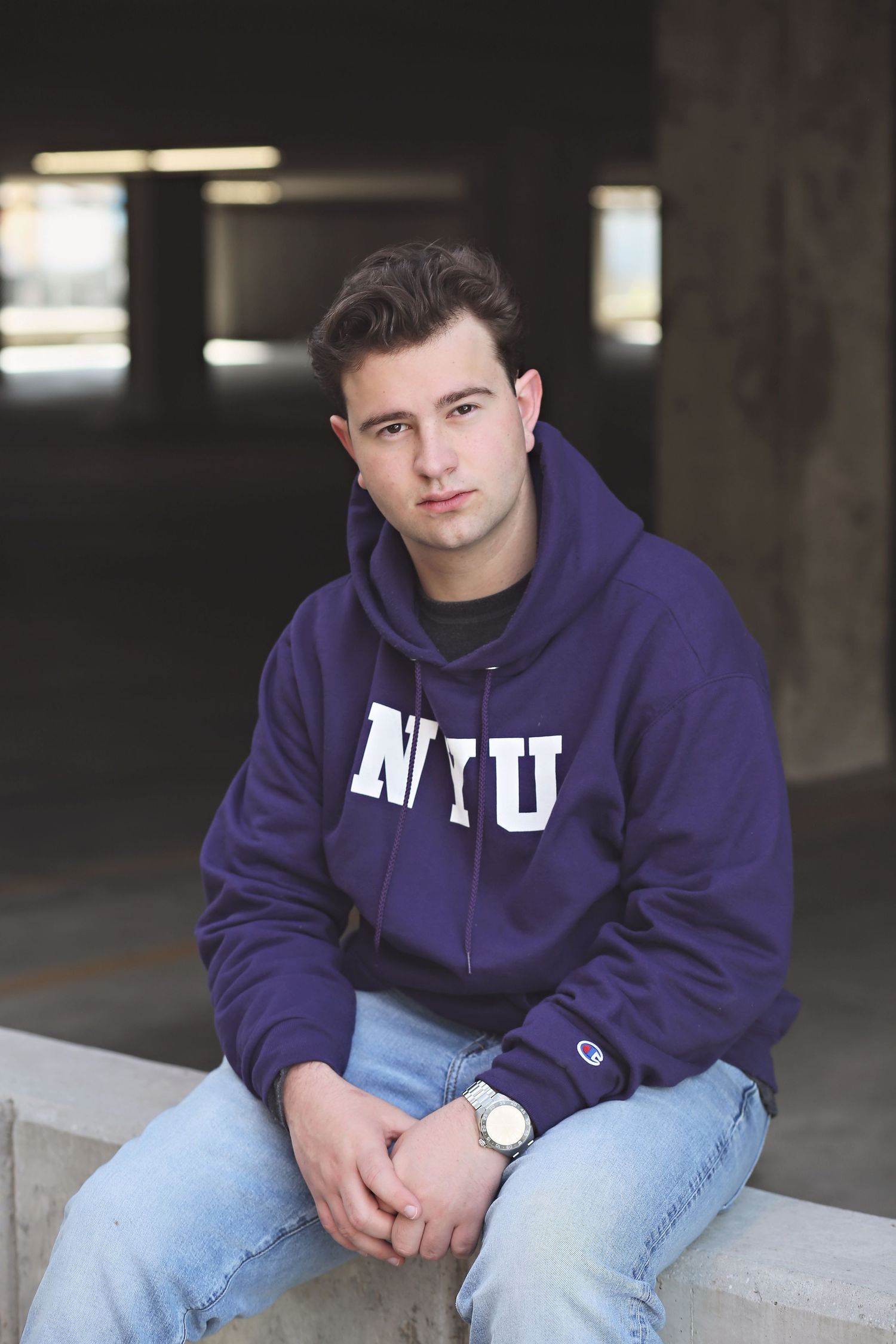 Person in purple NYU hoodie sits casually on concrete ledge in dark setting.