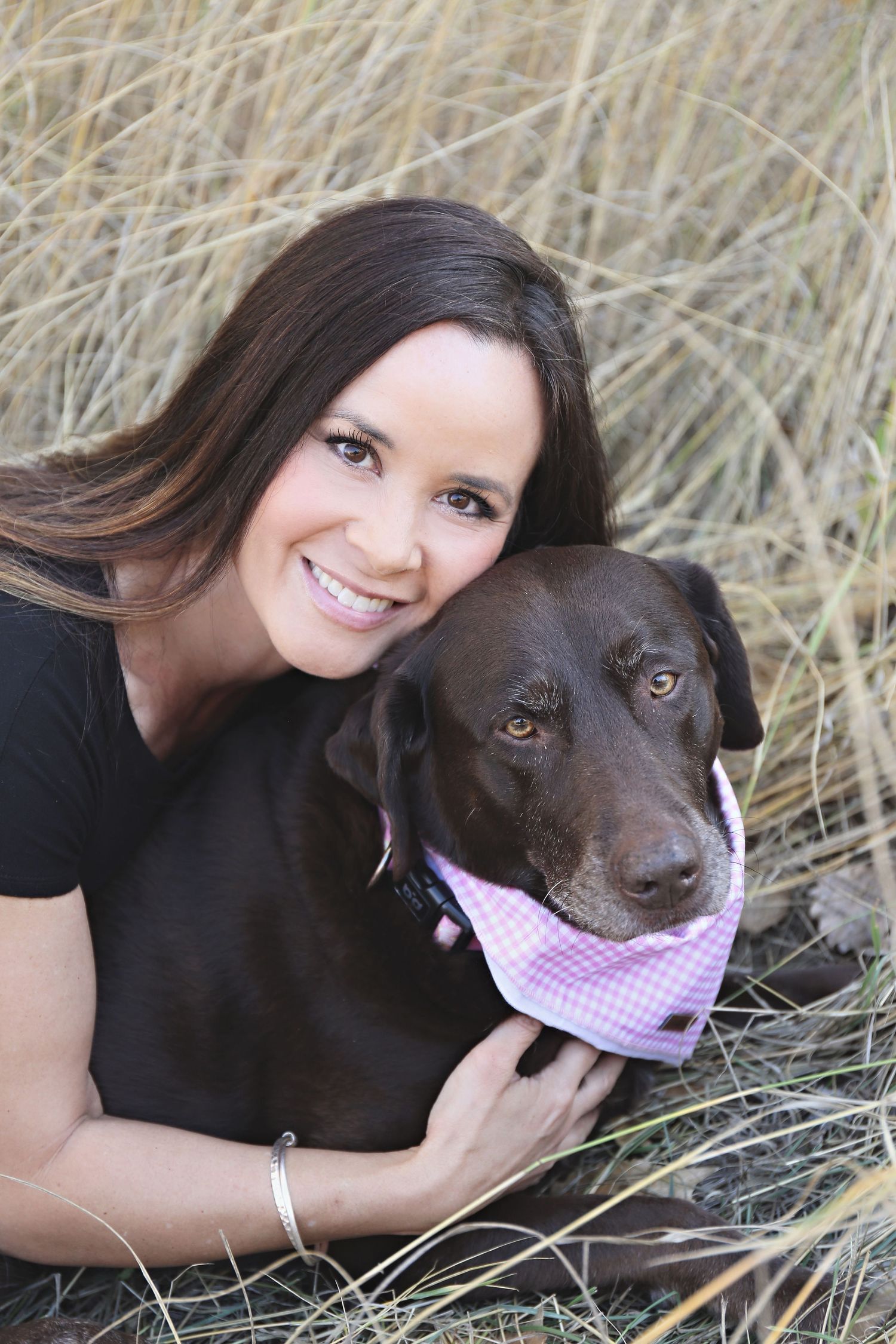 Chocolate lab wearing pink bandana poses with owner in tall grass field.