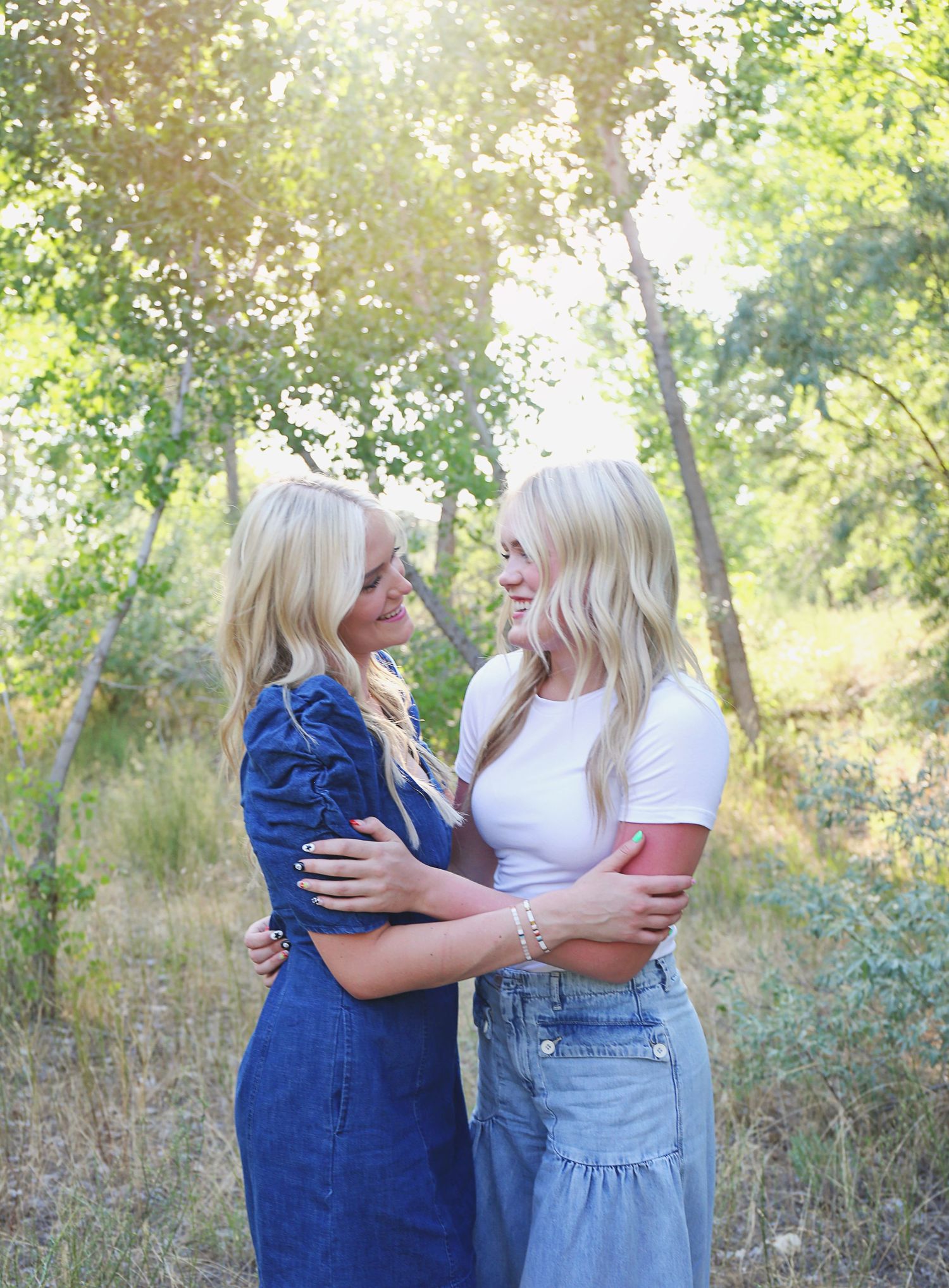 Two friends in denim dresses sharing a warm moment outdoors in natural lighting.