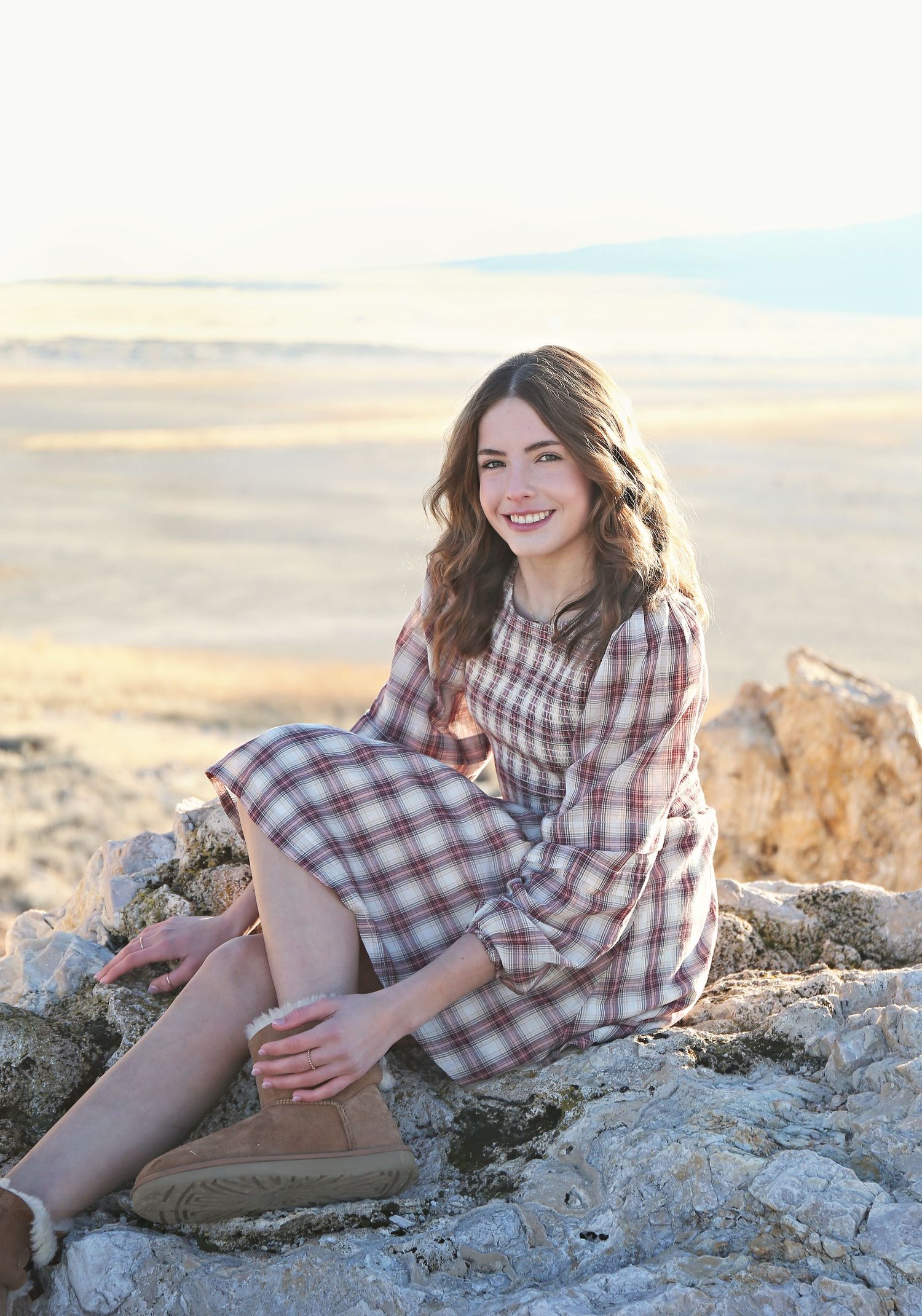 A woman in a checkered dress sits on rocks during sunset in a desert landscape.