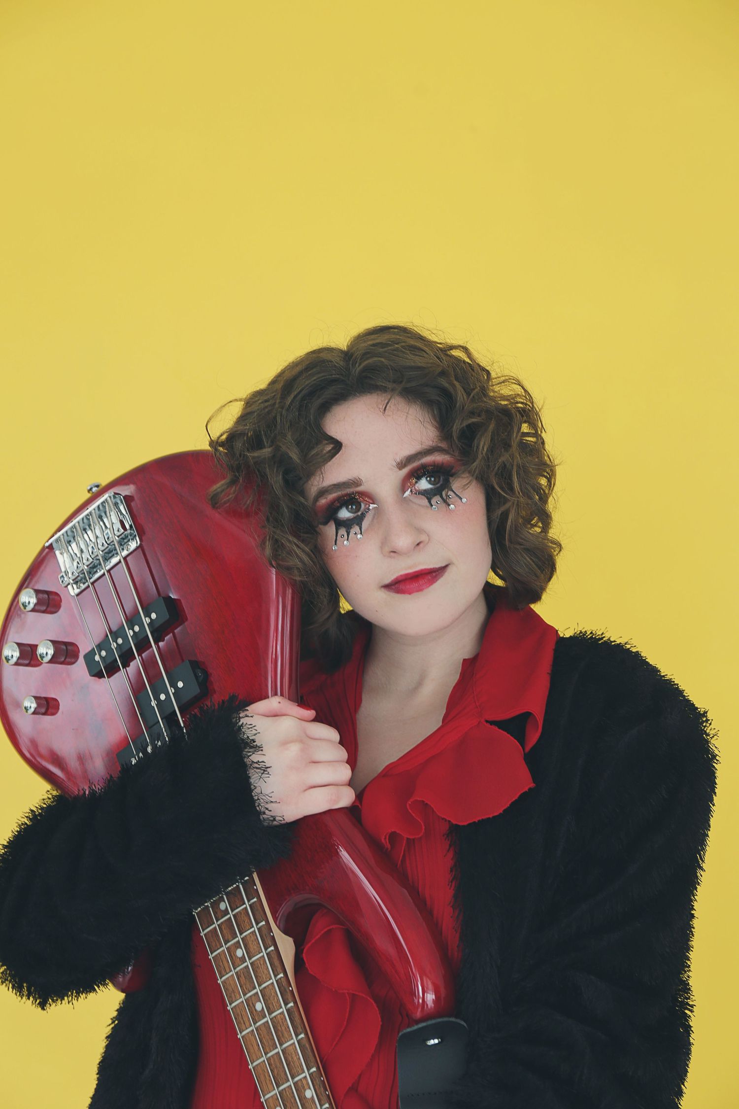 A musician in a black fur coat and red blouse poses with a red electric bass guitar against a yellow background.