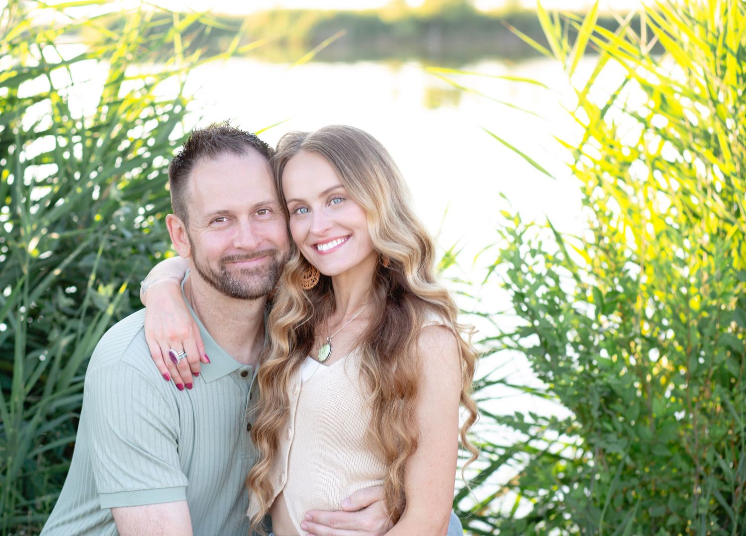 A couple poses together outdoors in front of green foliage on a sunny day.