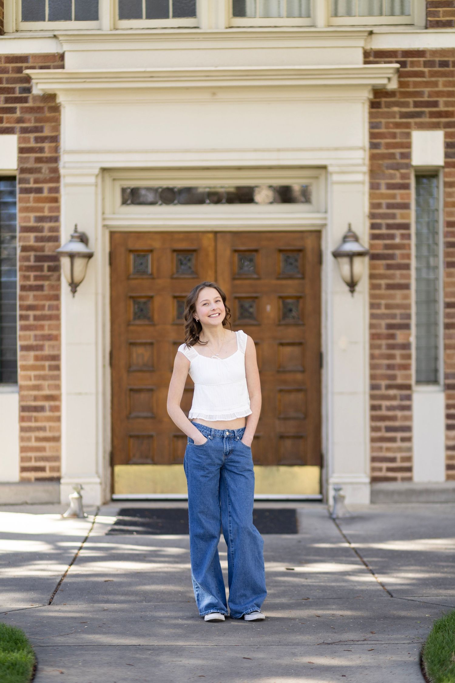 Young woman in white tank top and wide-leg jeans smiling in front of a brick building with wooden doors.