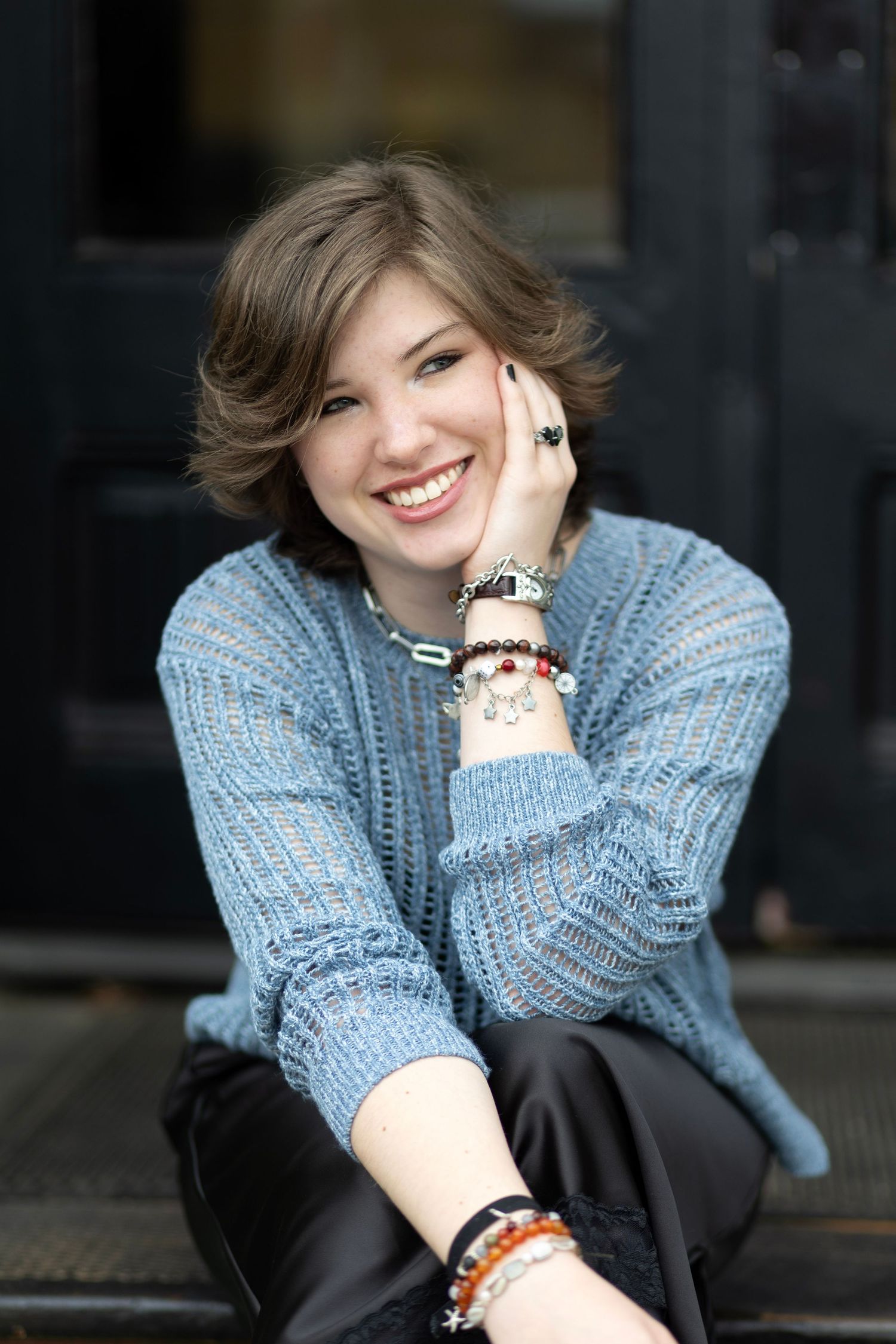 Smiling young woman in blue knit sweater with bracelets, sitting outdoors with hand on cheek.