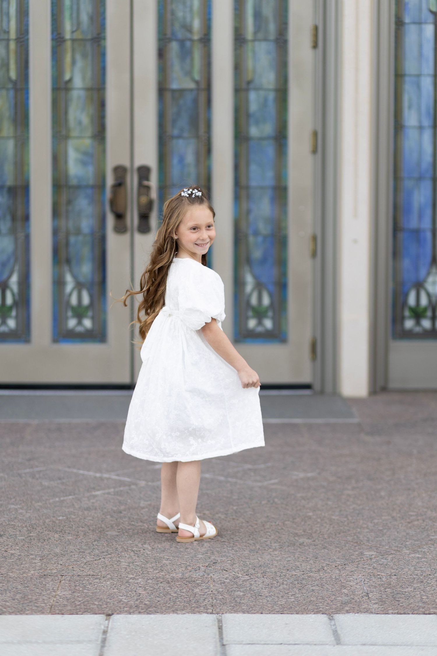 Young girl in white dress and hair crown smiles over shoulder near stained glass church doors.