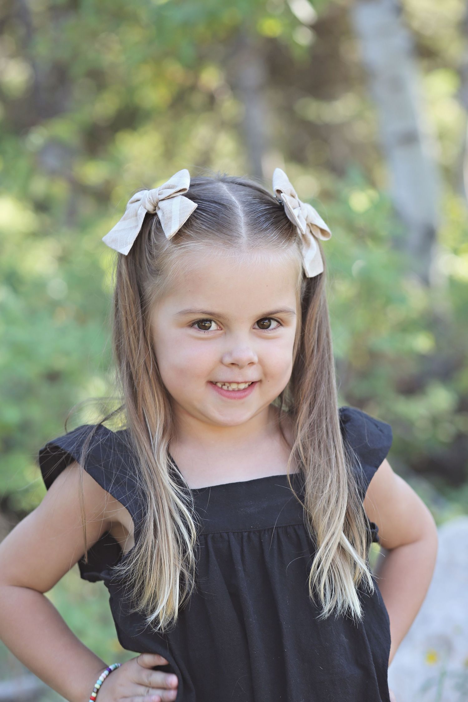 Young child in black dress with white hair bows poses outdoors for portrait series.