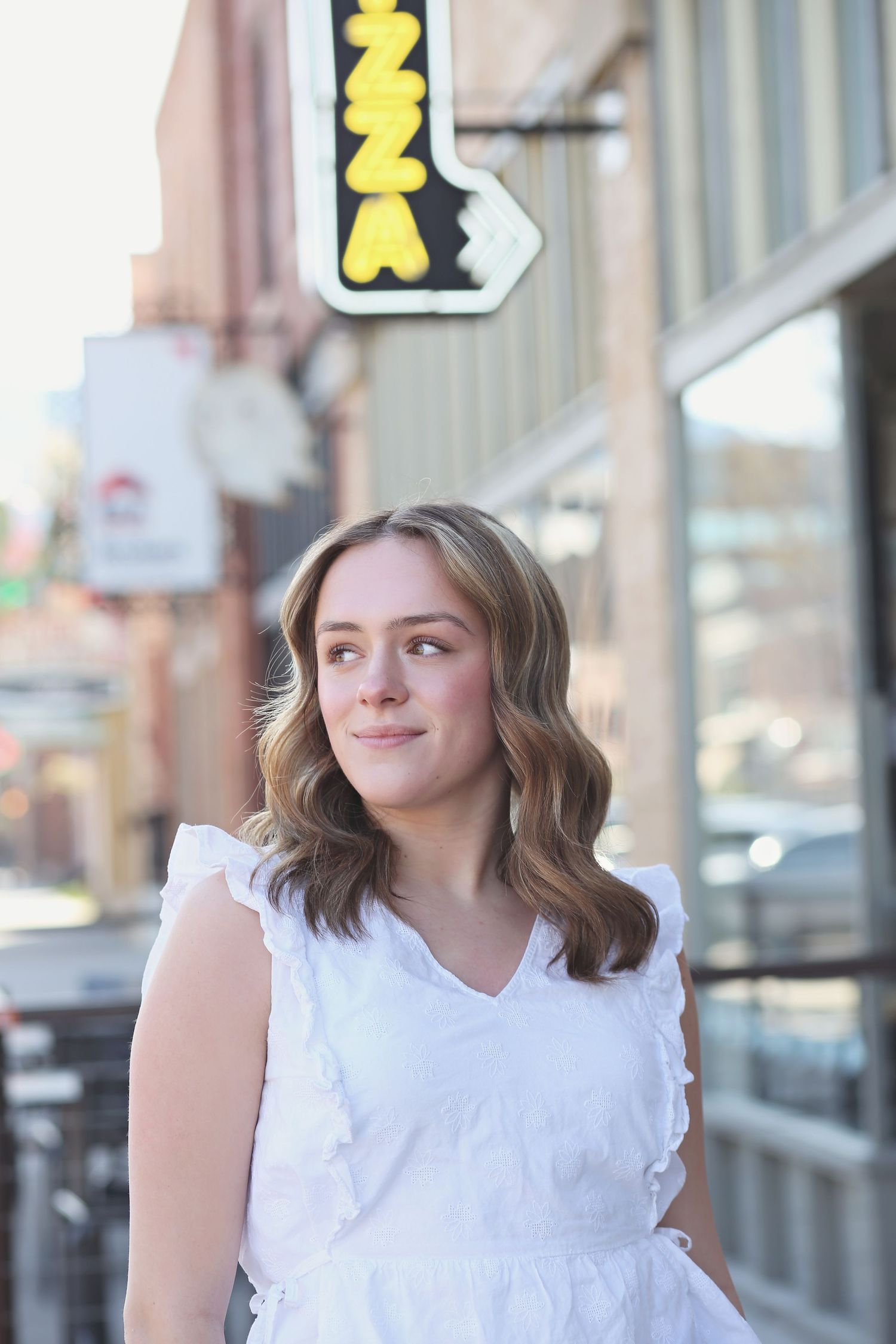 Young person in white ruffled top posing on urban street with illuminated sign in background.