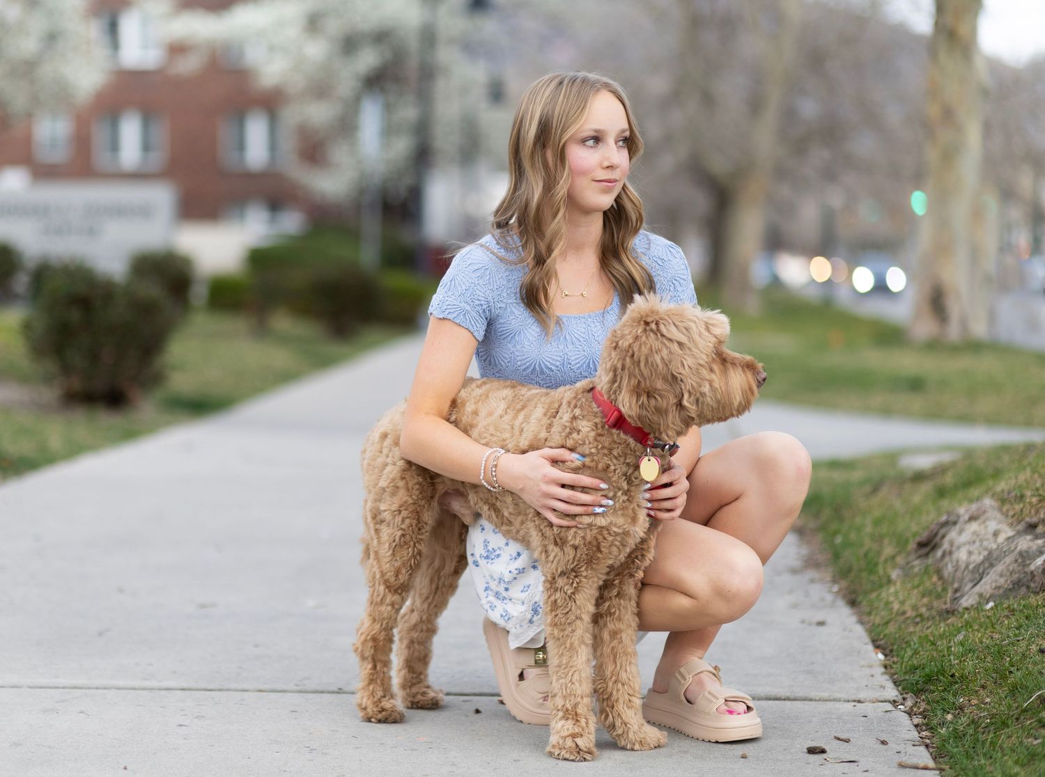Young woman kneeling on sidewalk hugging her fluffy golden doodle dog with red collar in park.