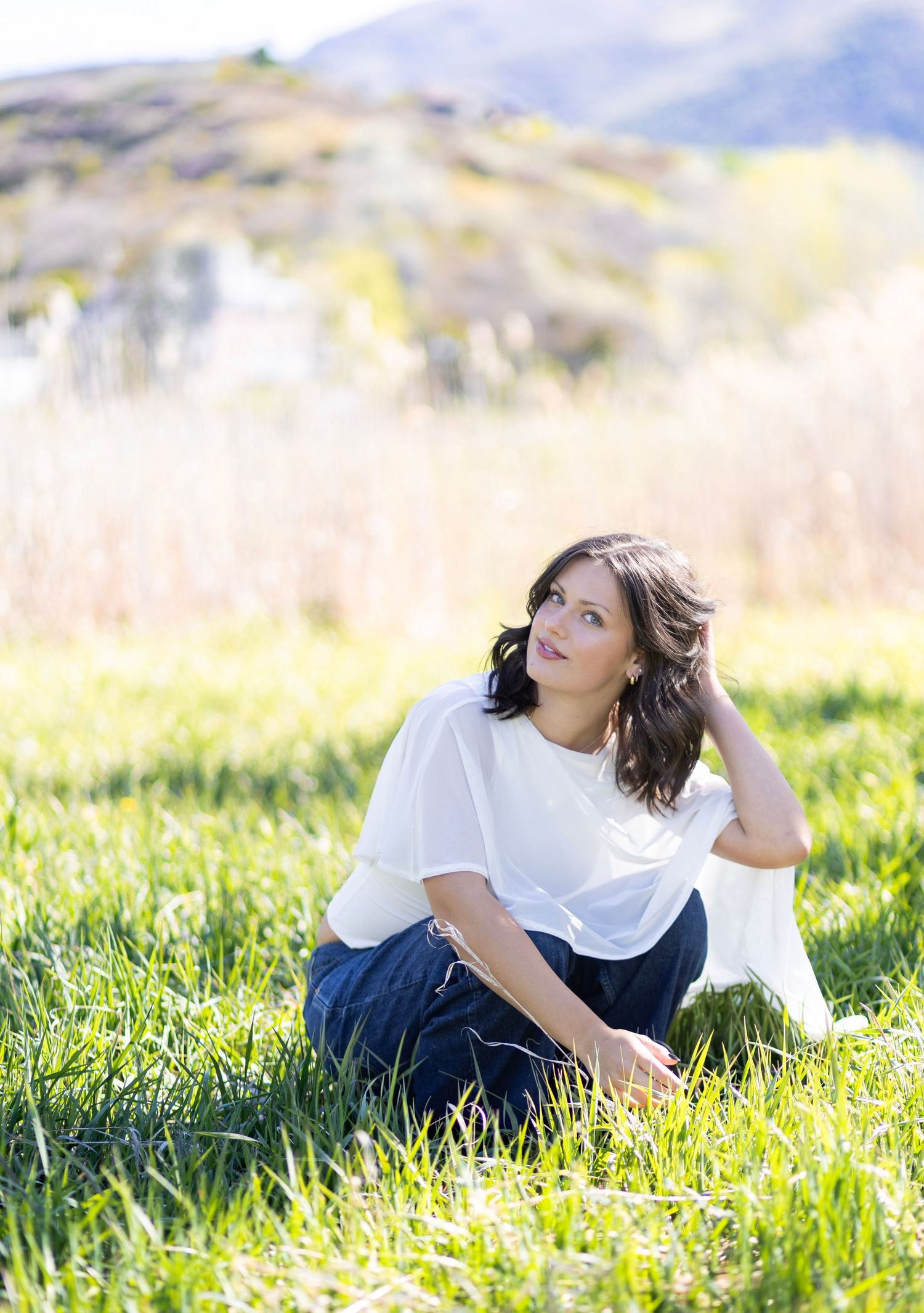 Woman in white top and jeans sitting in green grass field with mountain backdrop on sunny day.