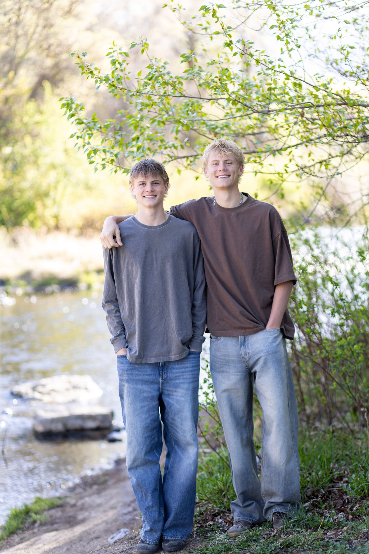 Two smiling teenage boys stand together near a creek outdoors, wearing casual t-shirts and jeans in a natural setting.