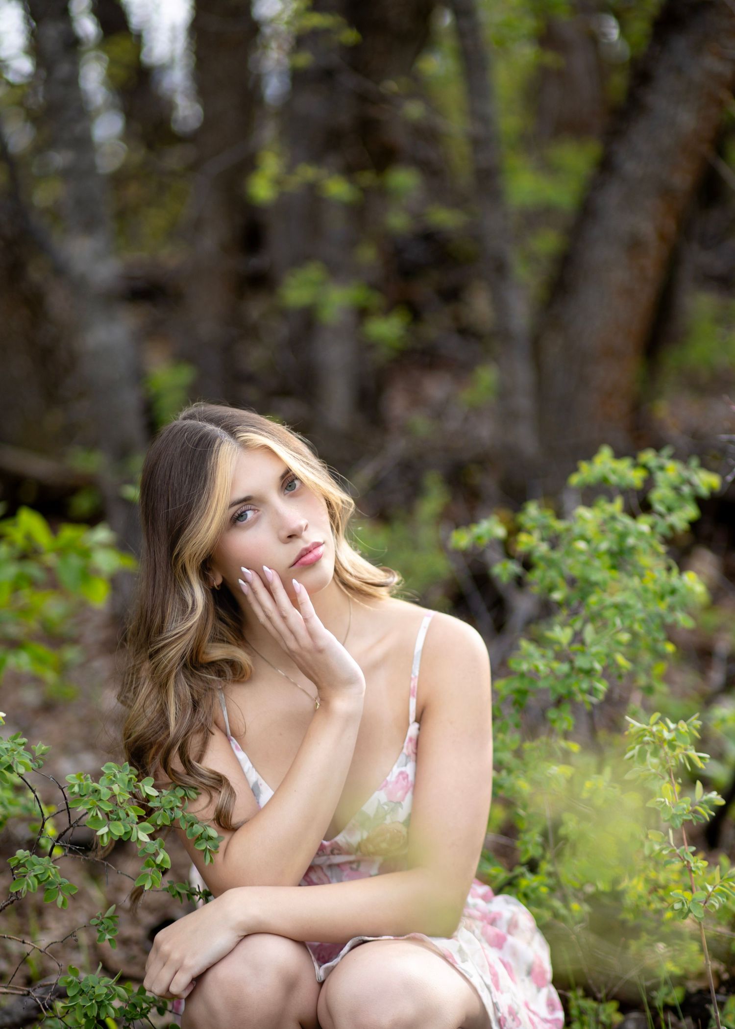 Young woman in floral dress sitting thoughtfully among green shrubs in a forest setting.