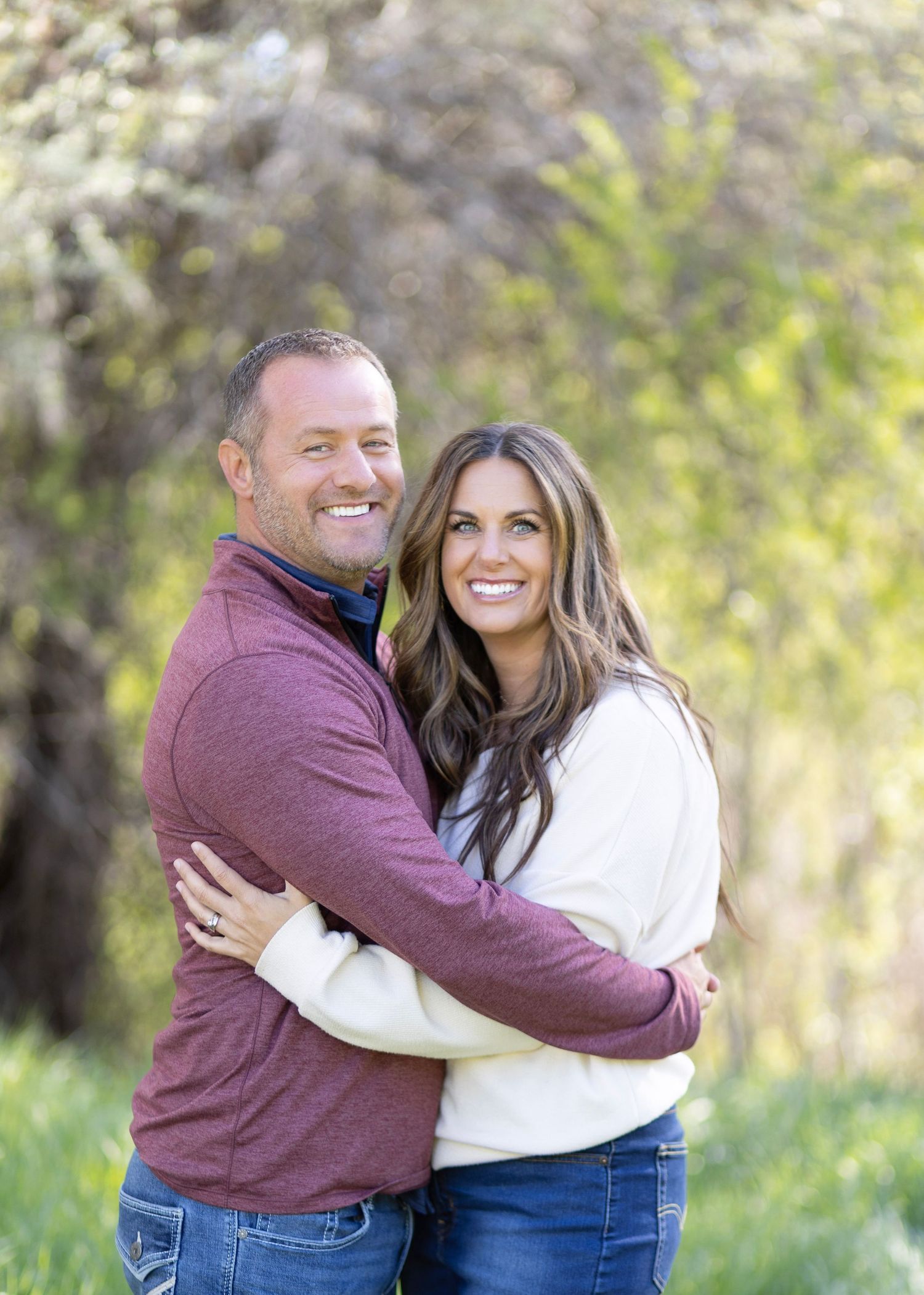 A smiling couple embraces outdoors in front of lush green trees, wearing casual attire in a bright spring setting.