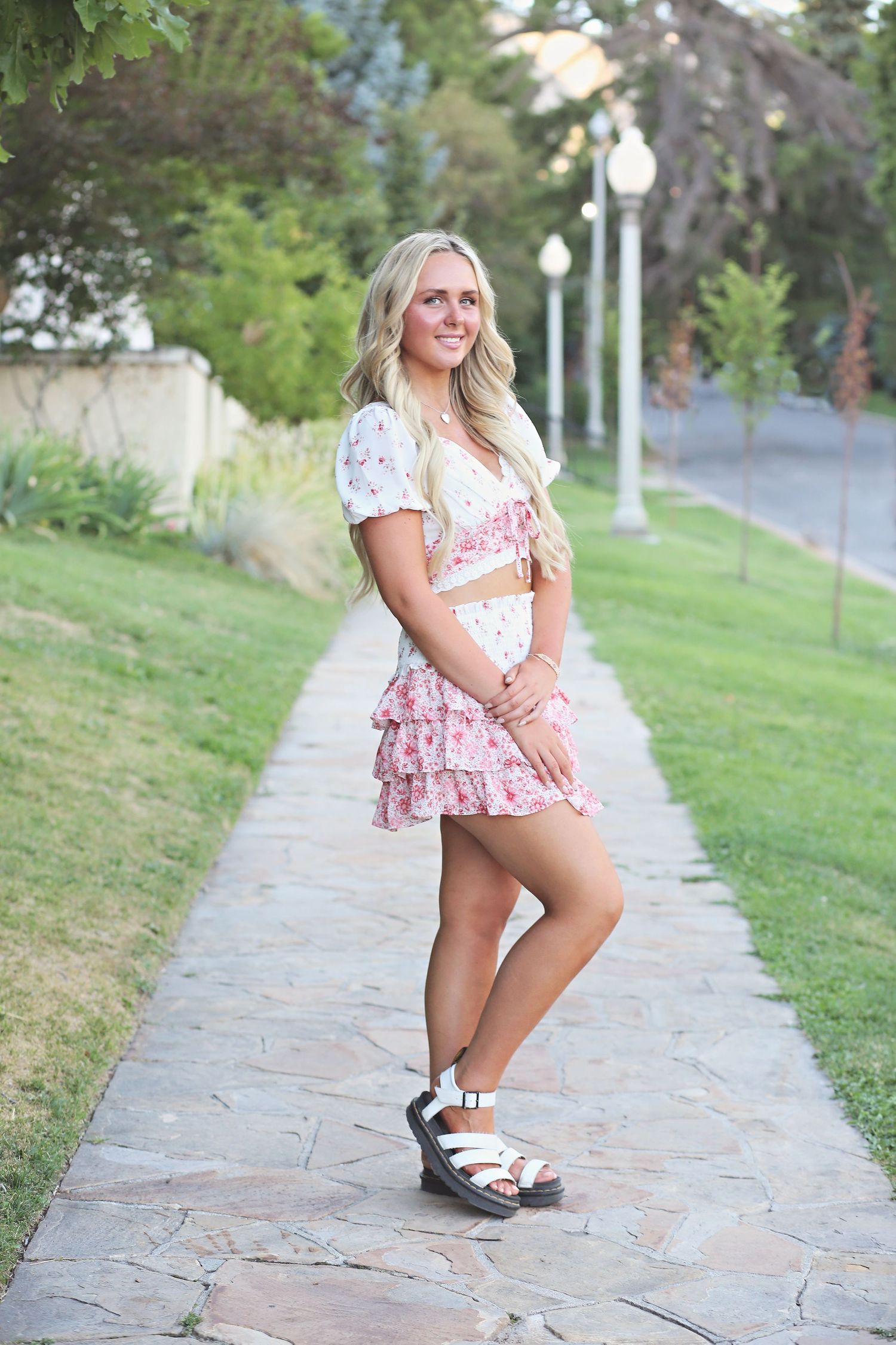 Person in floral dress poses on stone pathway surrounded by green landscaping.