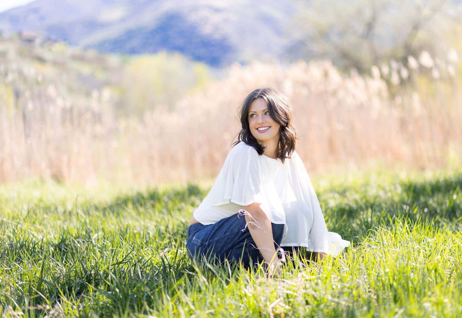 Woman sitting in green grass field smiling, wearing white top and blue skirt, with hills and tall grass in background.
