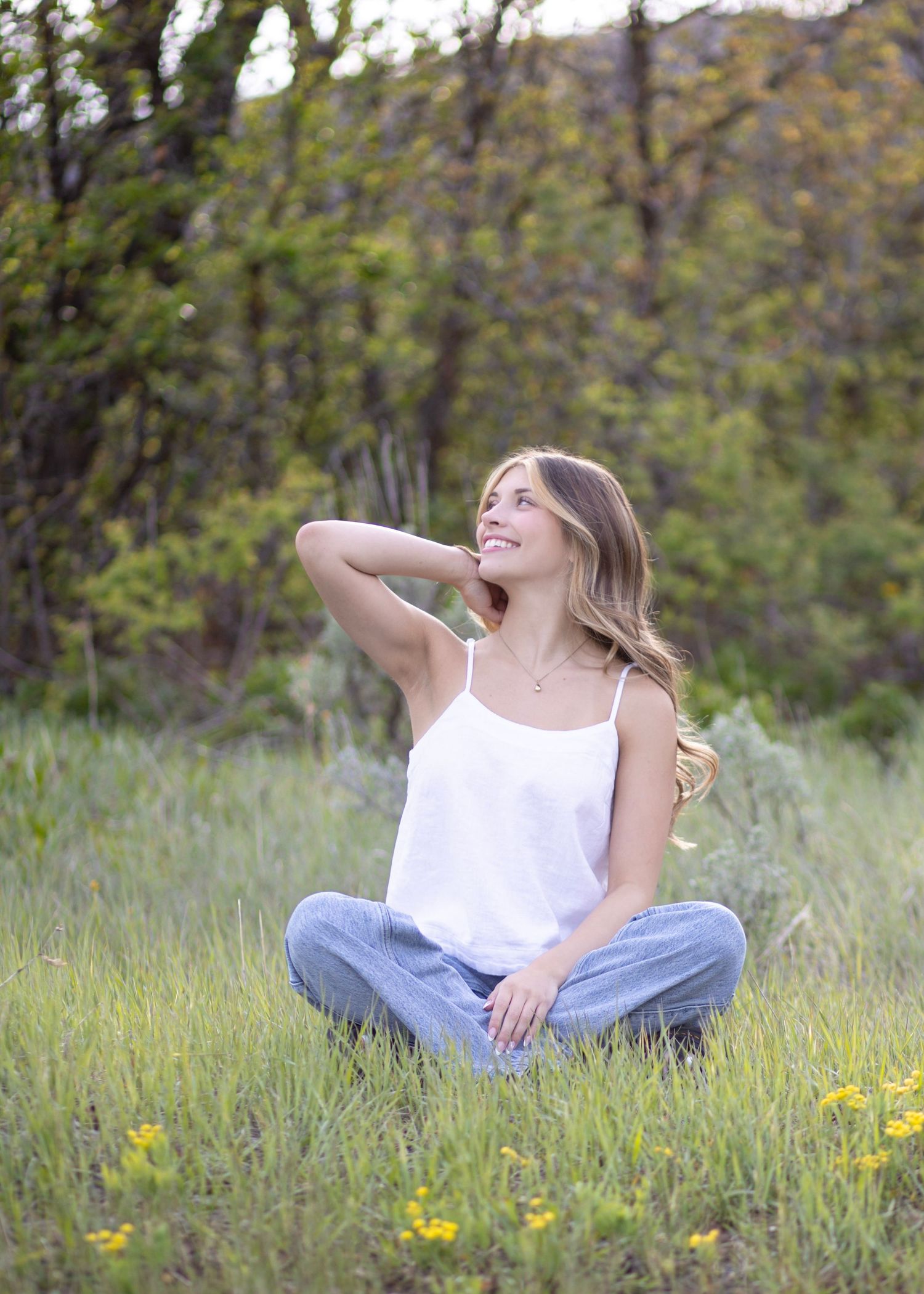 Smiling young woman in white tank and jeans sitting cross-legged in a lush green meadow with wildflowers.