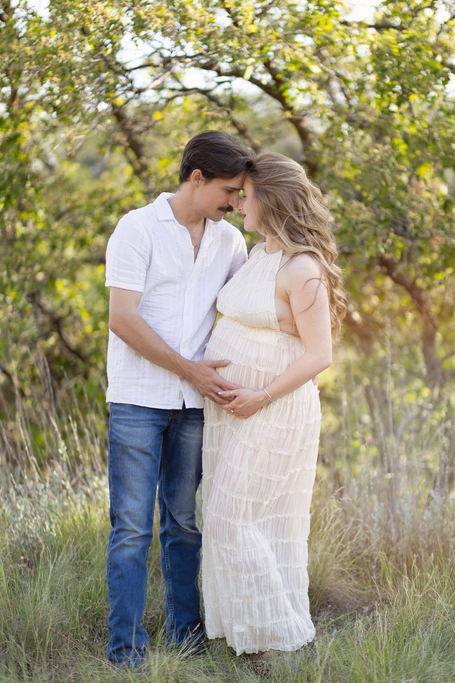 A couple tenderly embraces outdoors, the woman in a white dress showing a baby bump, surrounded by lush green trees.