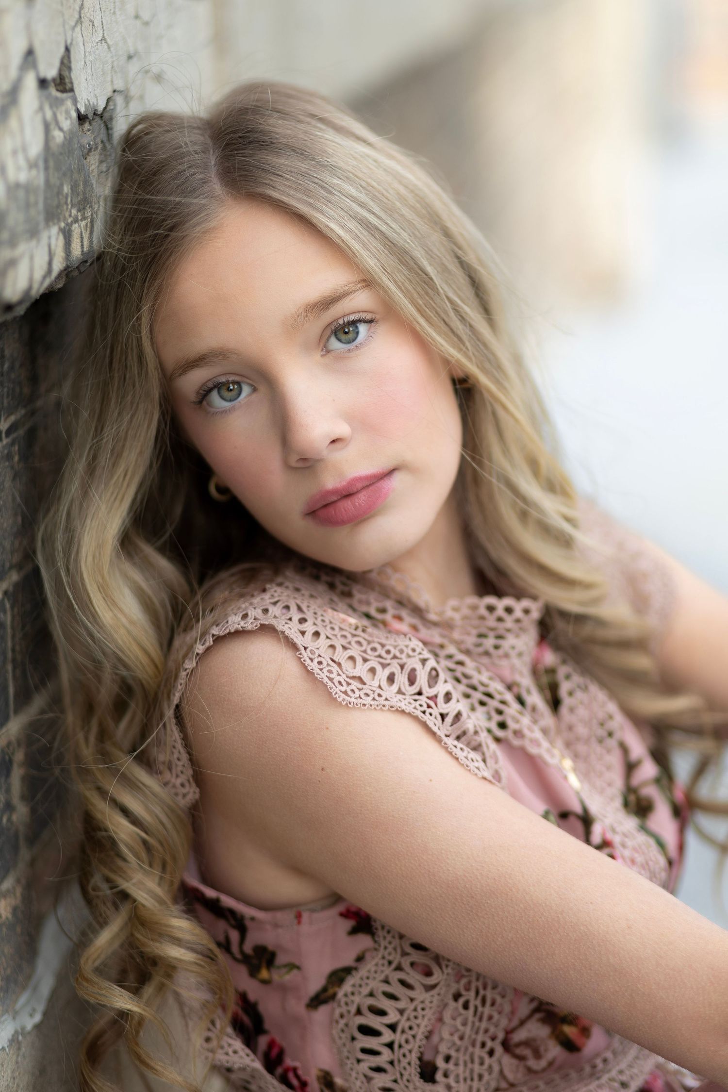 A young girl with long blonde wavy hair leans against a stone wall wearing a pink floral lace dress.