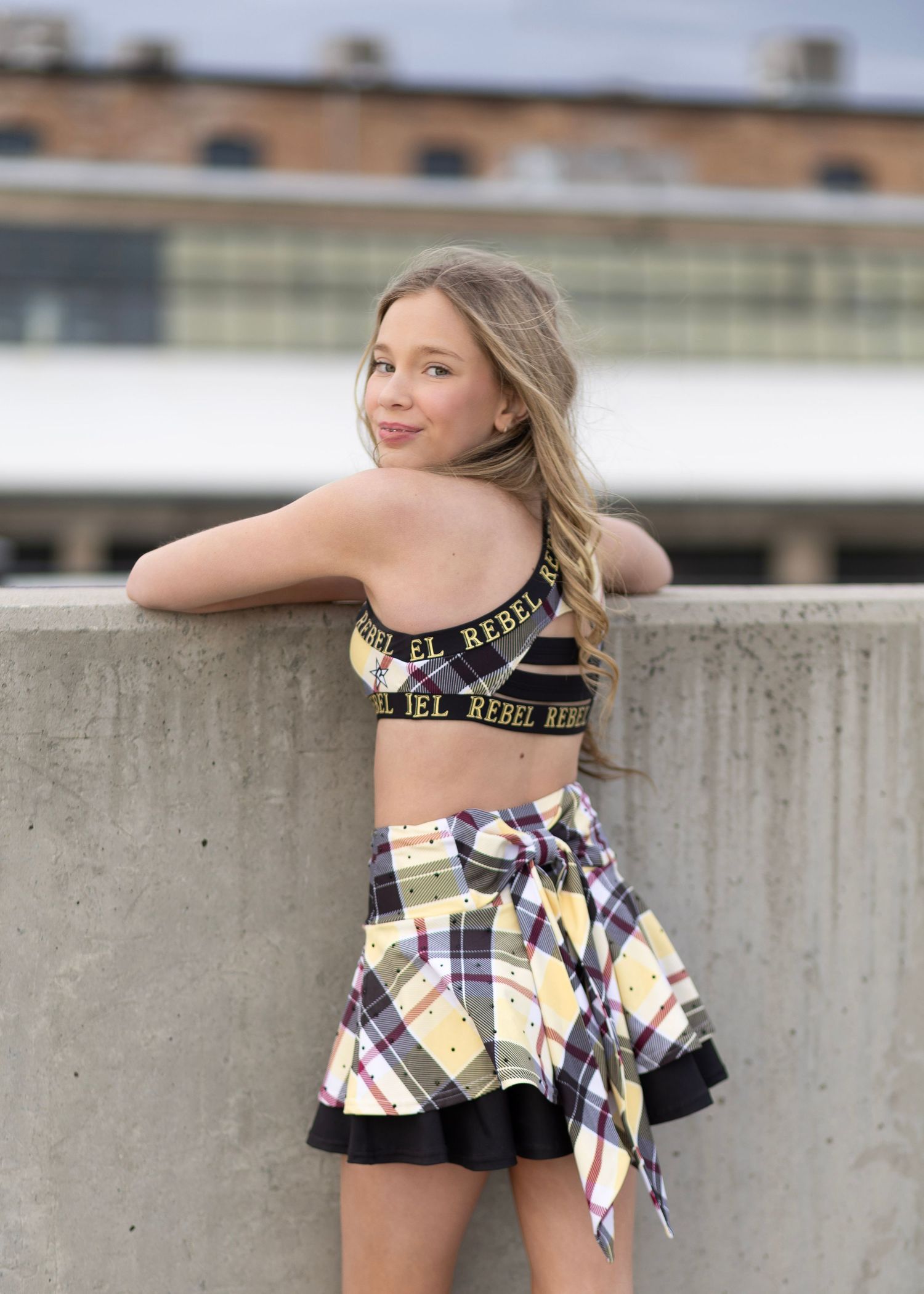 Young girl in a plaid skirt and black rebel crop top posing outdoors on a rooftop ledge.