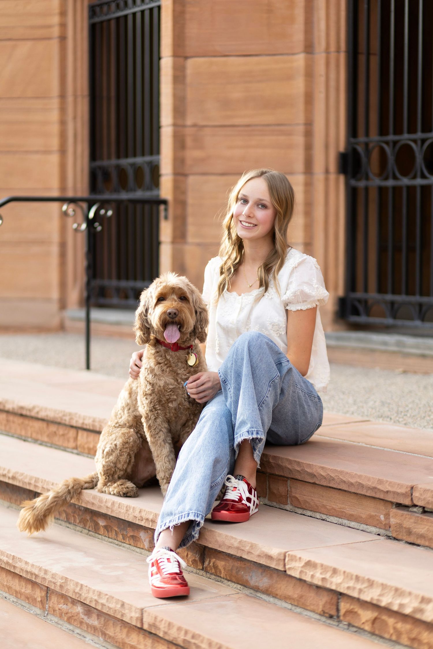 Smiling blonde woman in white top and jeans sitting on brick steps with her brown Labradoodle dog.