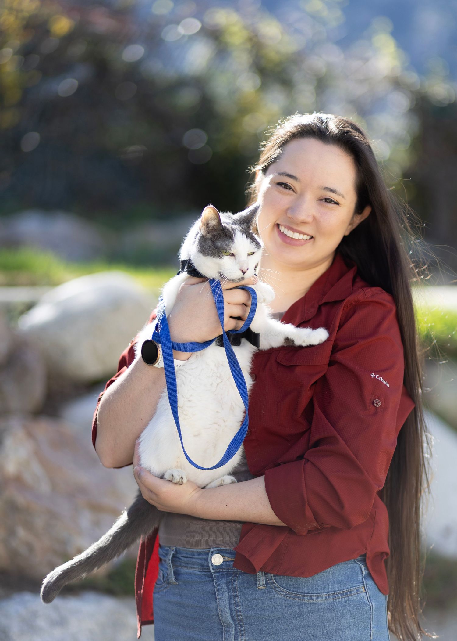 Smiling woman in red jacket holding gray and white cat on blue leash outdoors.