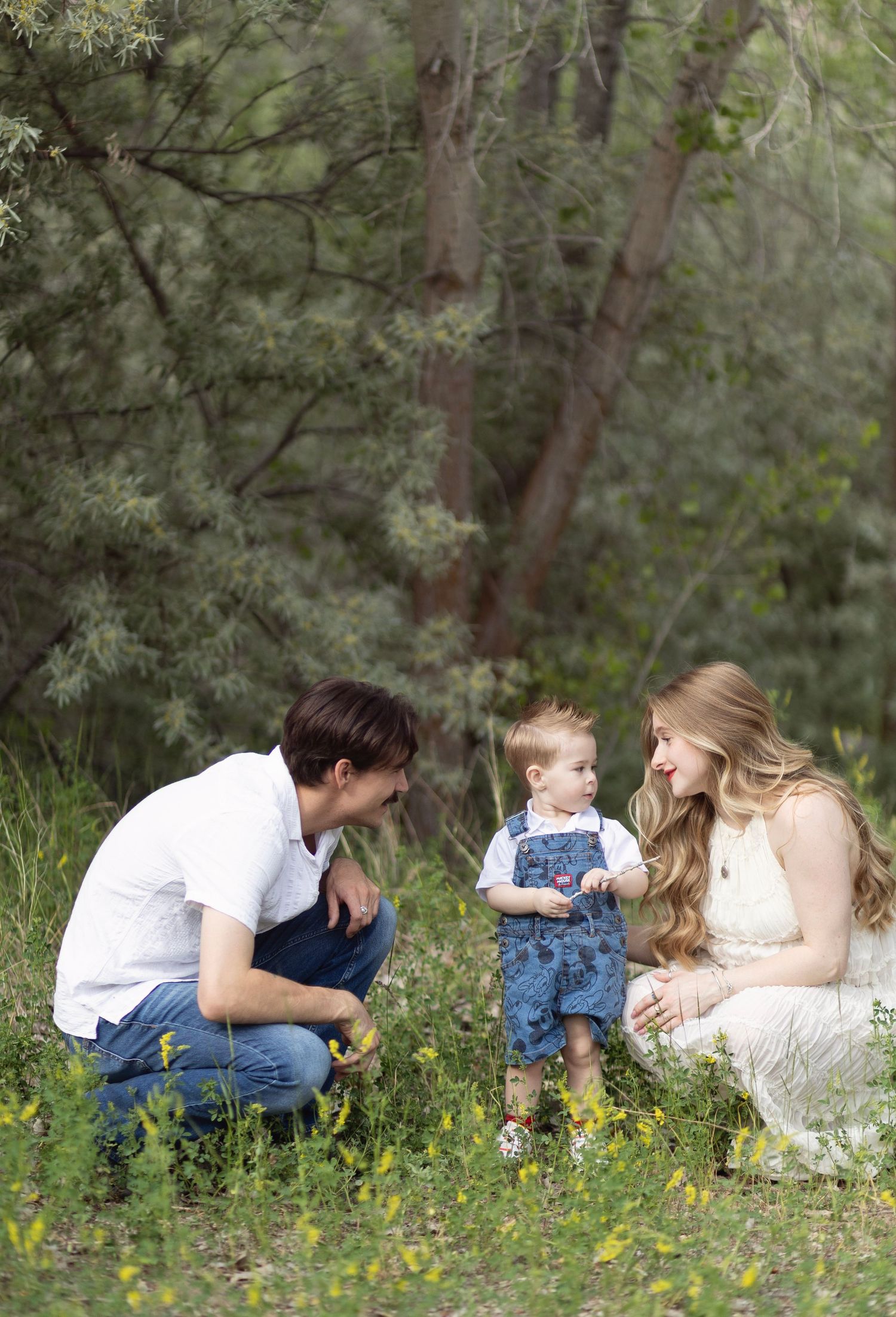 Family of three with toddler boy in denim overalls sharing a sweet moment outdoors among wildflowers and trees.
