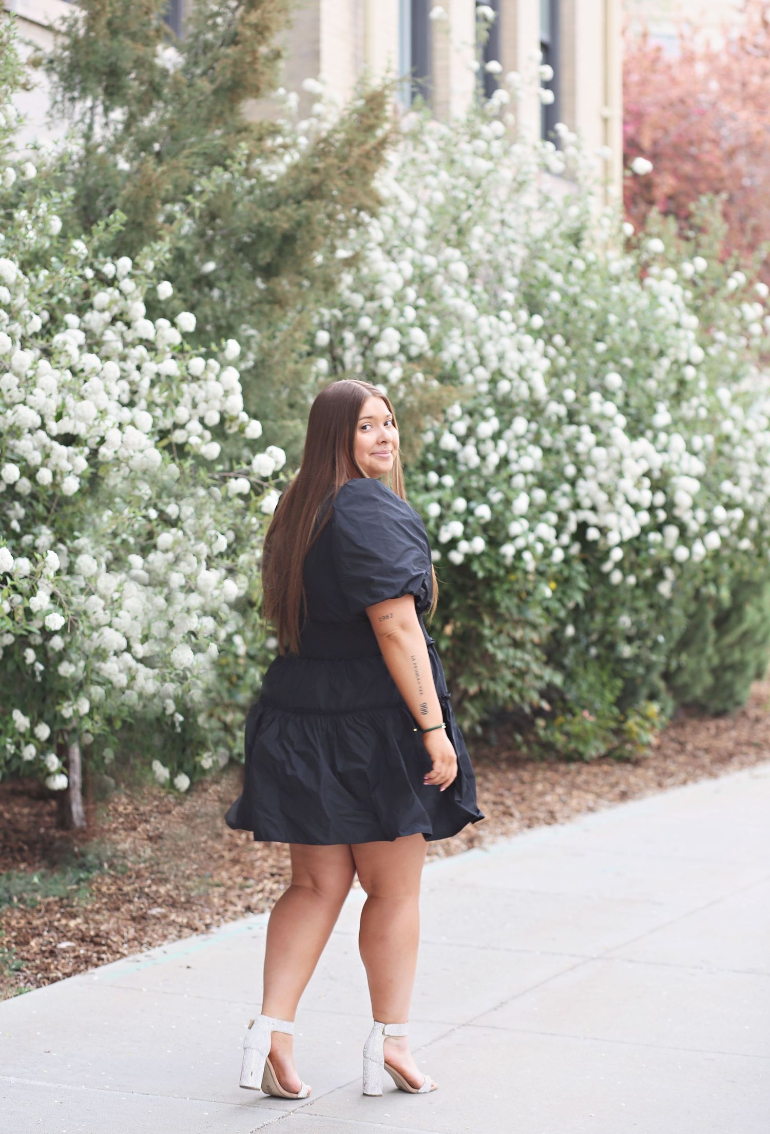 Graduate in black dress poses near blooming white flowers on campus walkway.