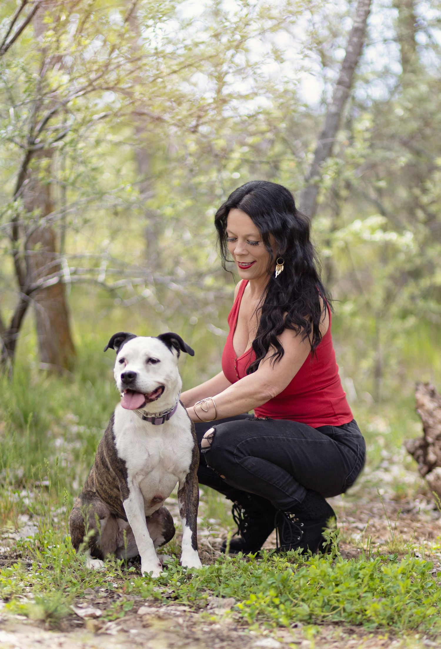 Woman in red top crouching beside a brindle pit bull dog in a lush green forest setting.
