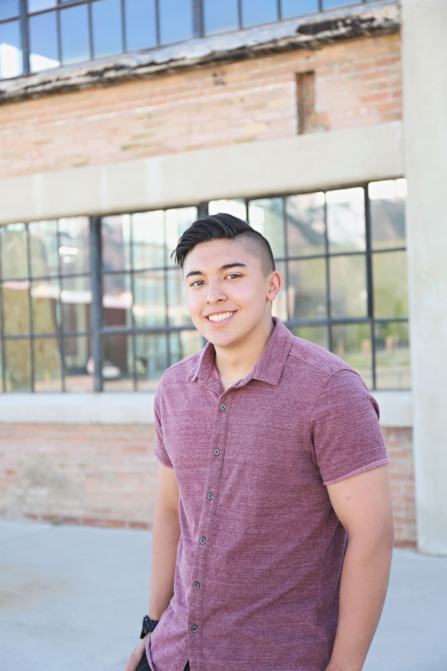 Young person in a burgundy polo shirt poses and smiles in front of a brick building with large windows.