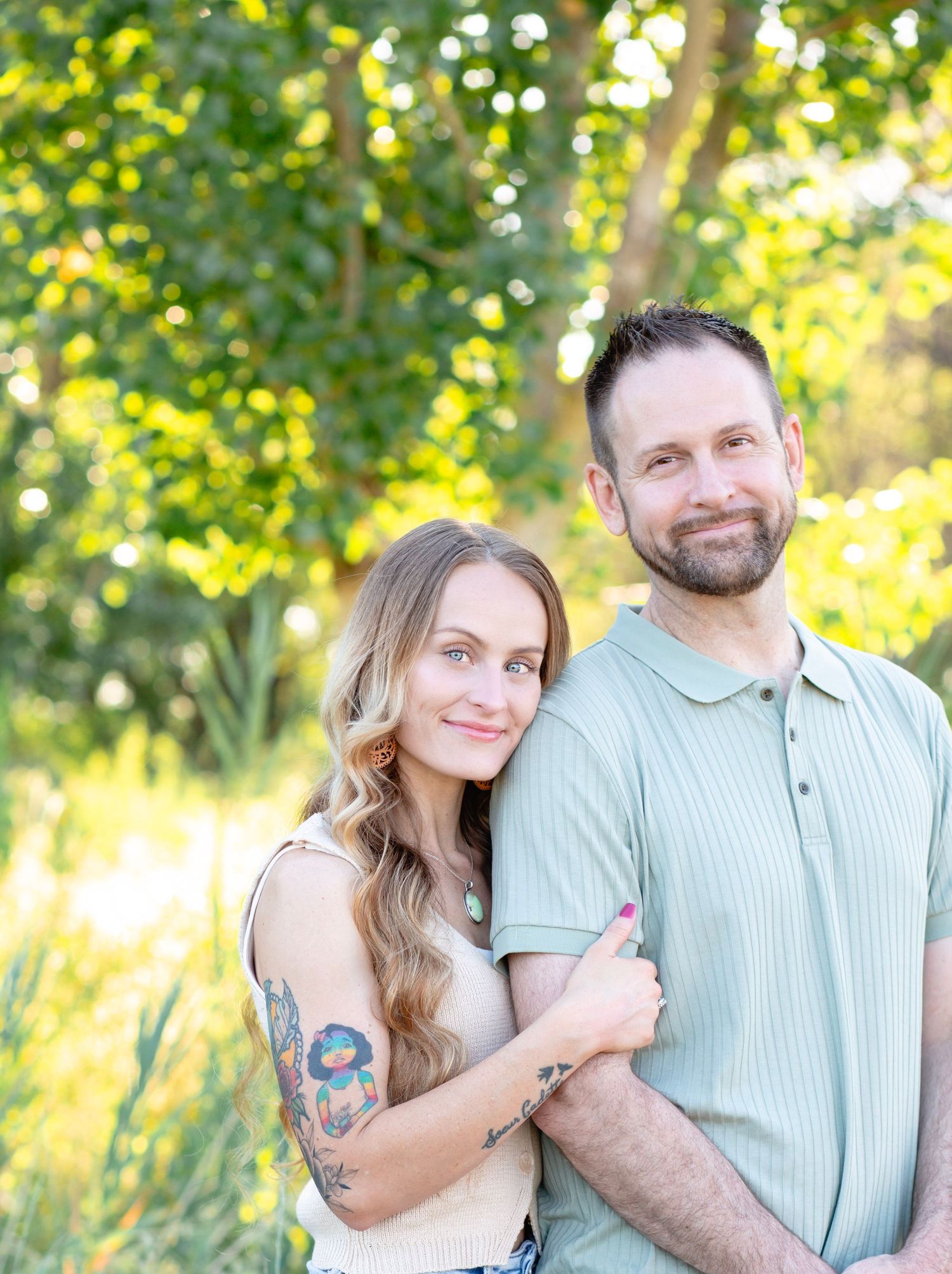 A couple poses together outdoors with green foliage and sunlight creating a romantic backdrop.