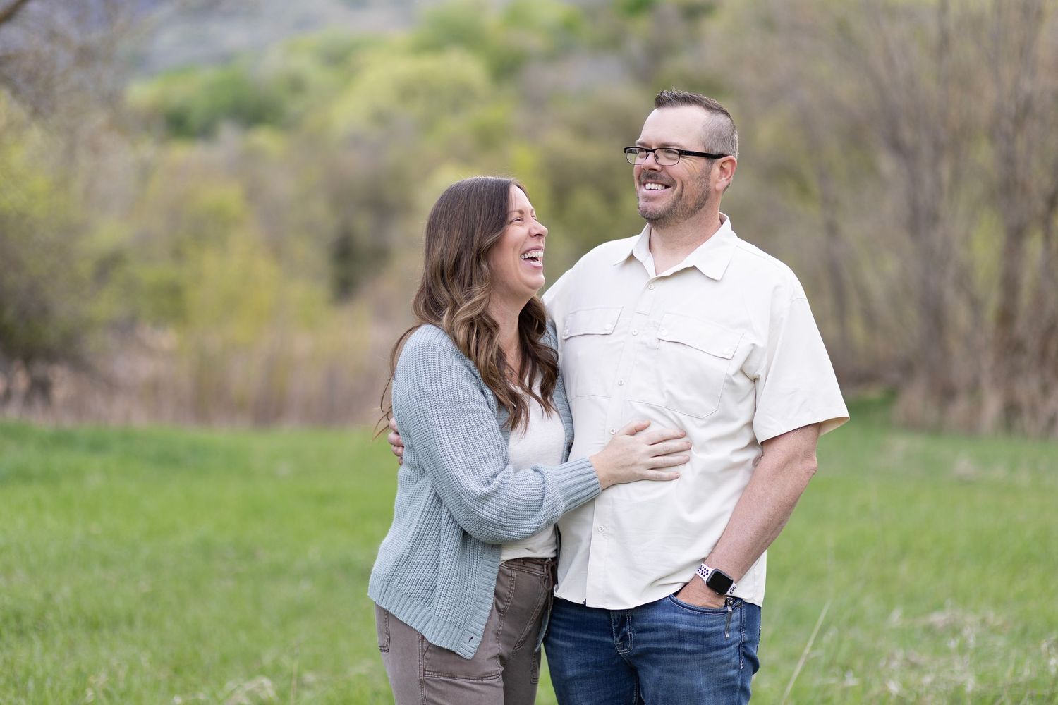 A laughing couple embraces outdoors in a green meadow, sharing a joyful moment during their engagement photo session.