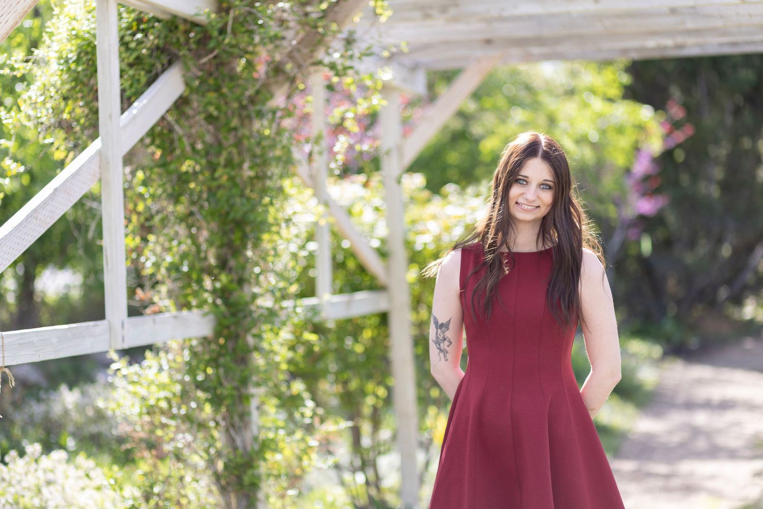Smiling brunette woman in burgundy dress standing under white pergola in garden.