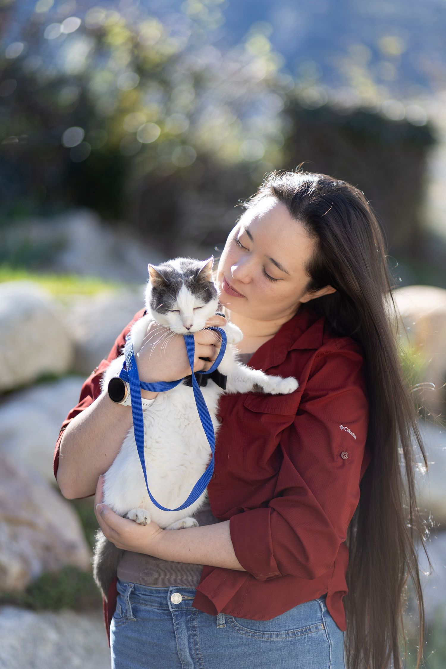Young woman in red shirt lovingly holding a gray and white cat on a blue leash outdoors.