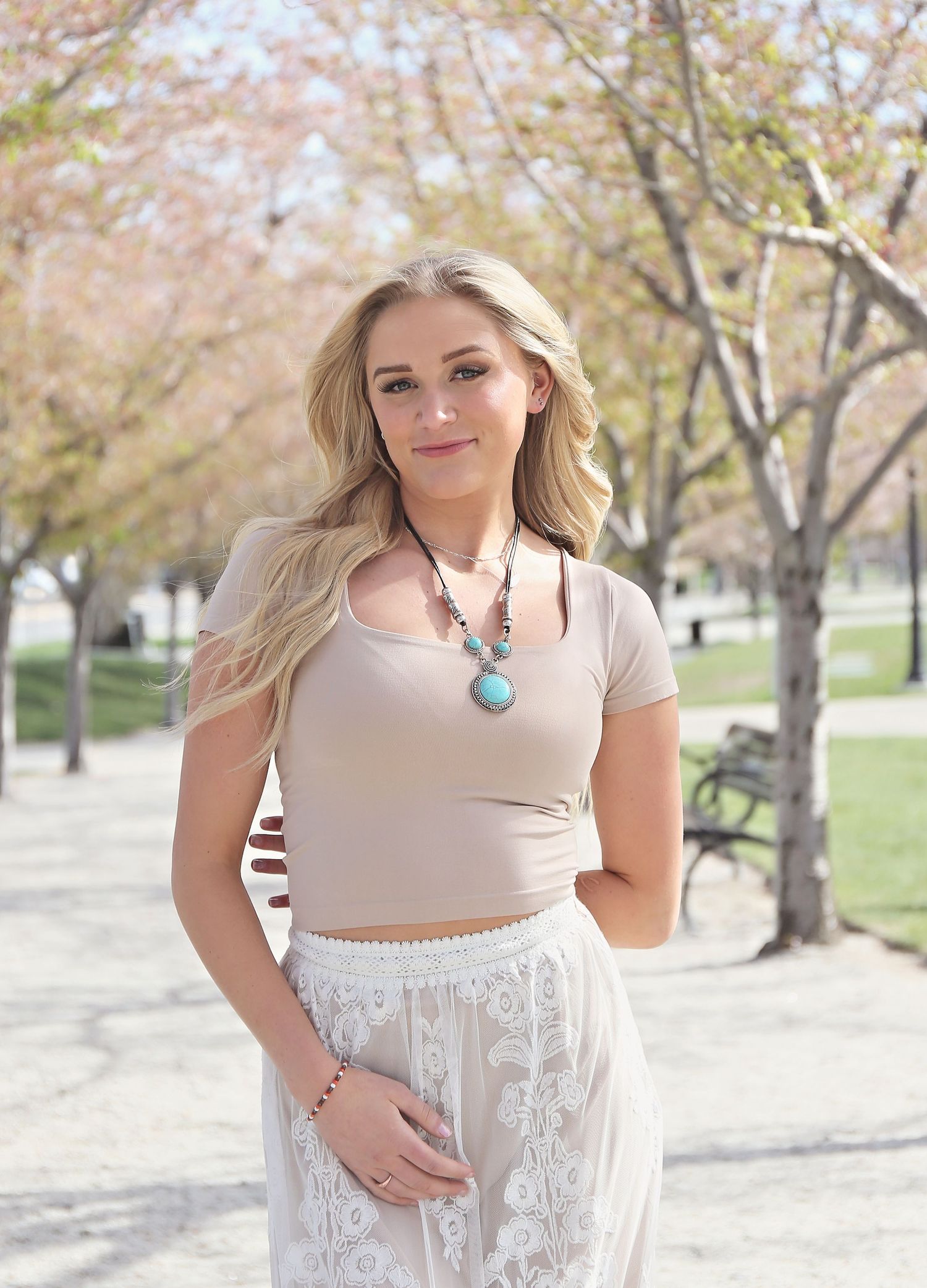 Person wearing beige crop top and white lace skirt posing in park with blooming trees.