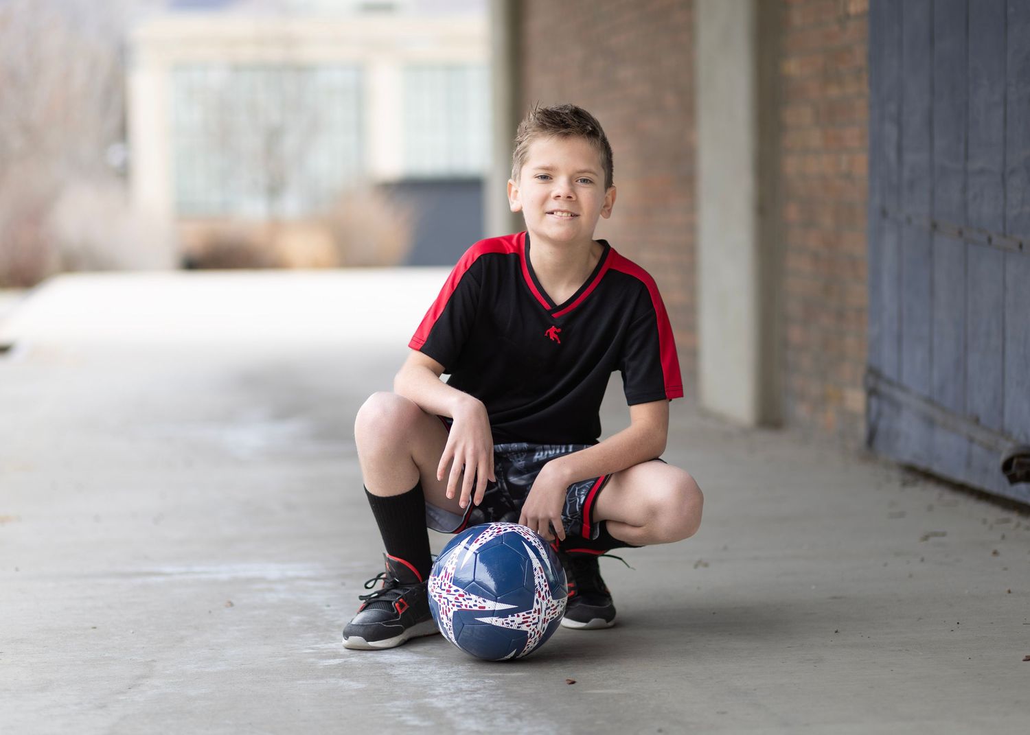 Young boy in black soccer uniform crouching with a soccer ball on pavement, smiling outdoors.