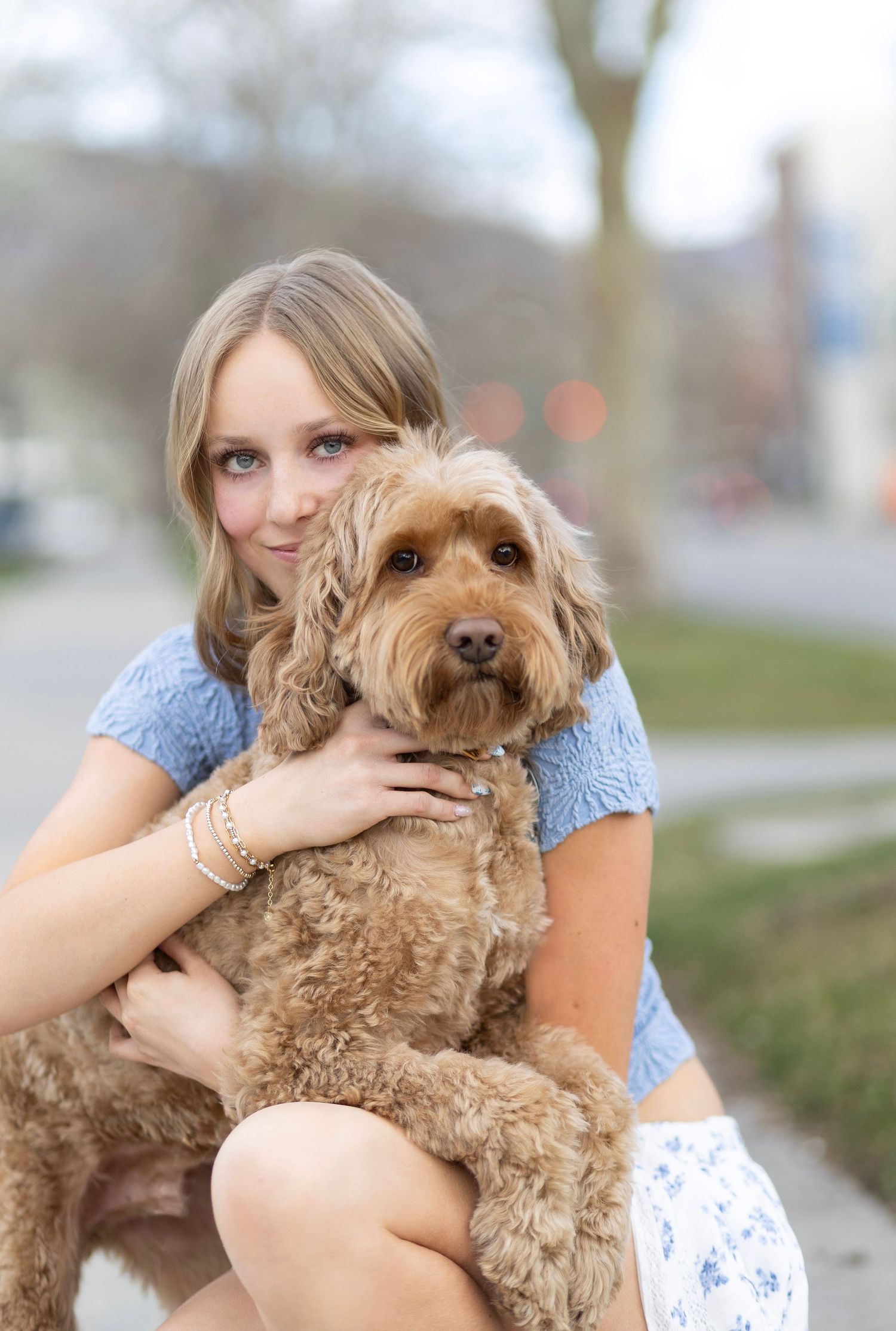 Young blonde woman in blue top hugging a fluffy golden doodle dog outdoors.