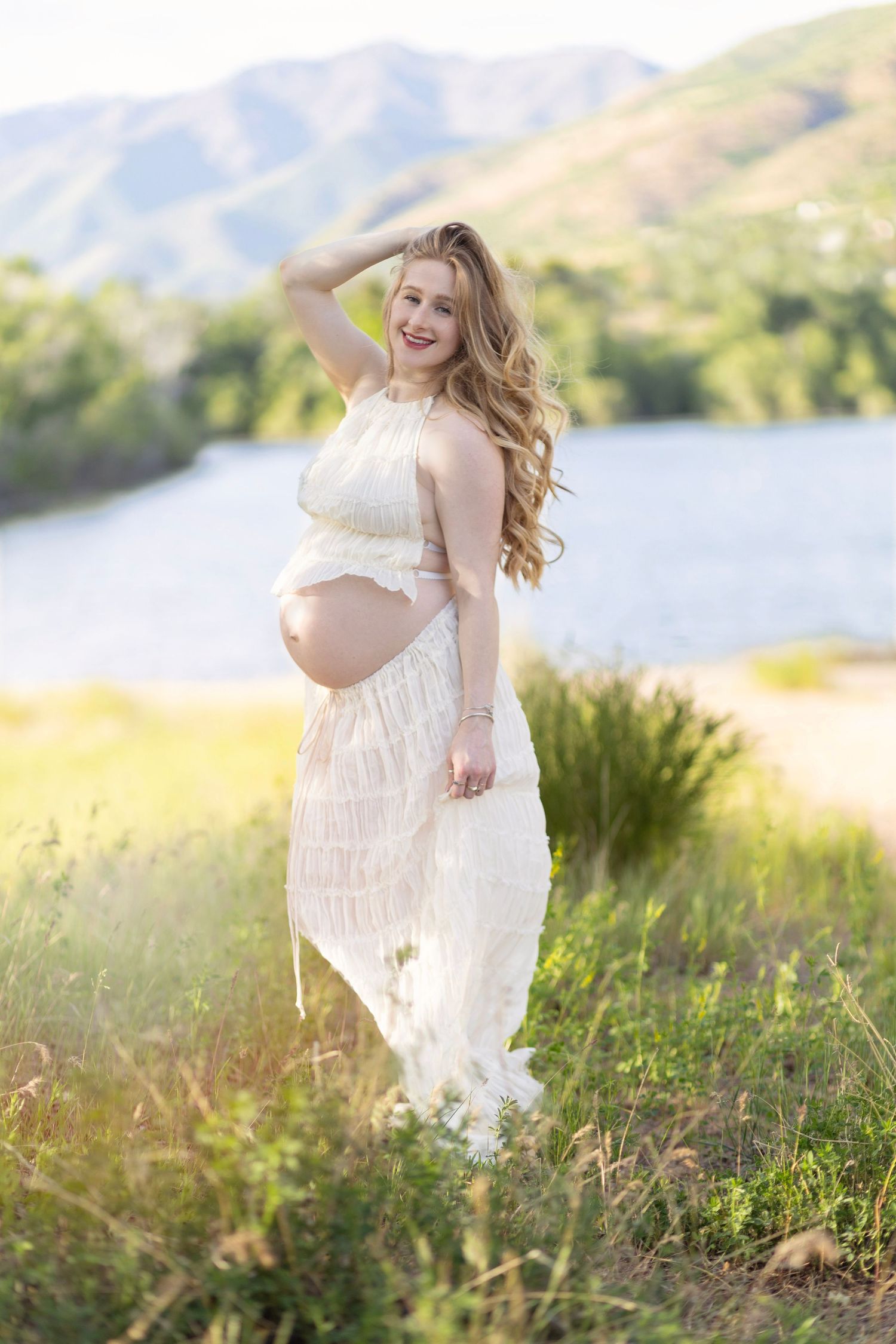 Smiling pregnant woman in white lace maternity dress posing outdoors near a lake with green hills in background.