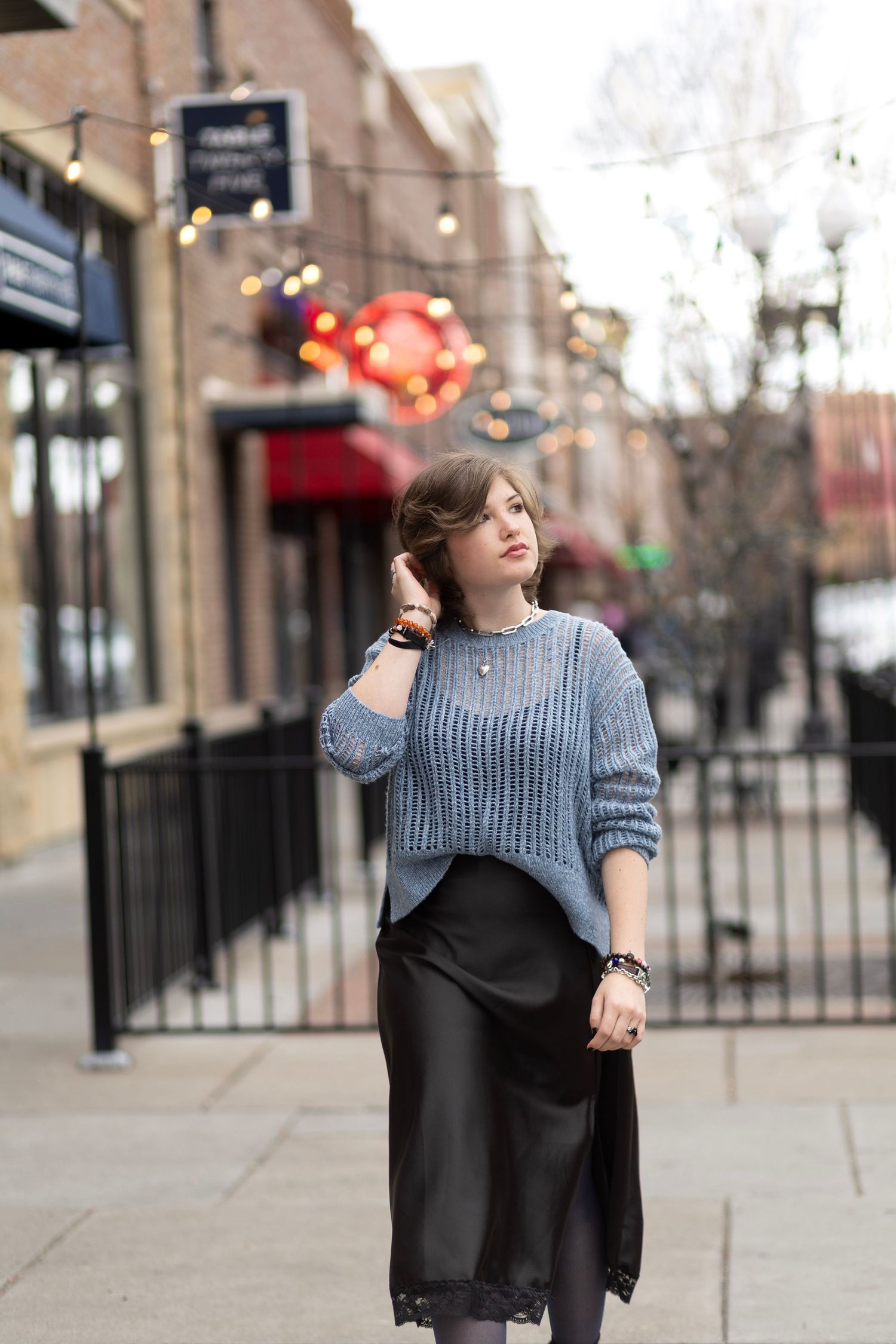 Woman in blue knit sweater and black satin skirt poses on urban sidewalk with bokeh string lights behind her.