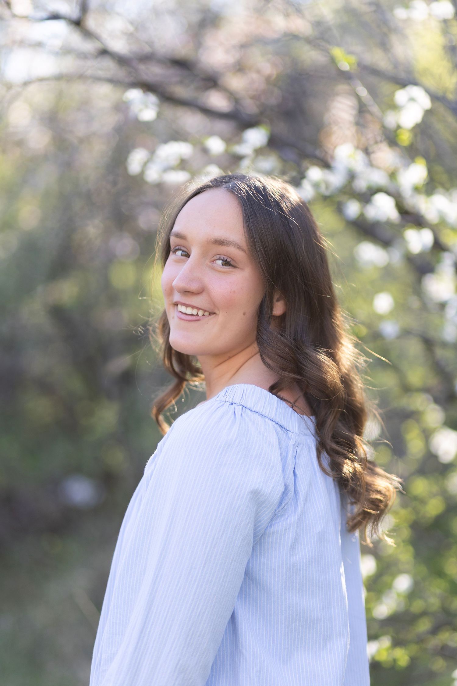 Young woman with long brown hair smiling outdoors in a light blue blouse with blooming trees in background.