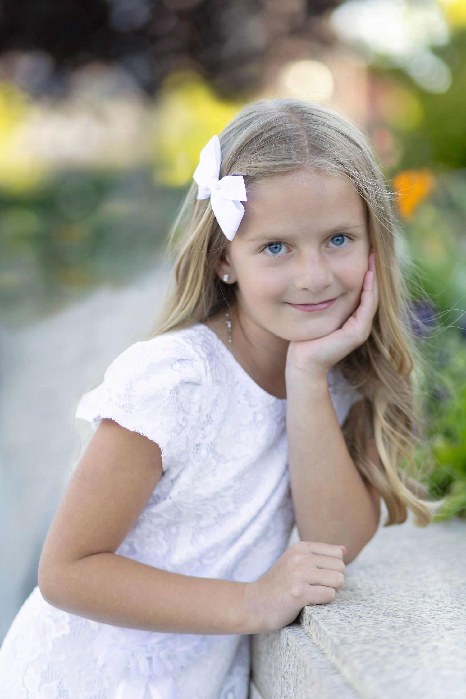 Young girl with blue eyes and blonde hair wearing a white dress and bow, posing outdoors in a garden setting.
