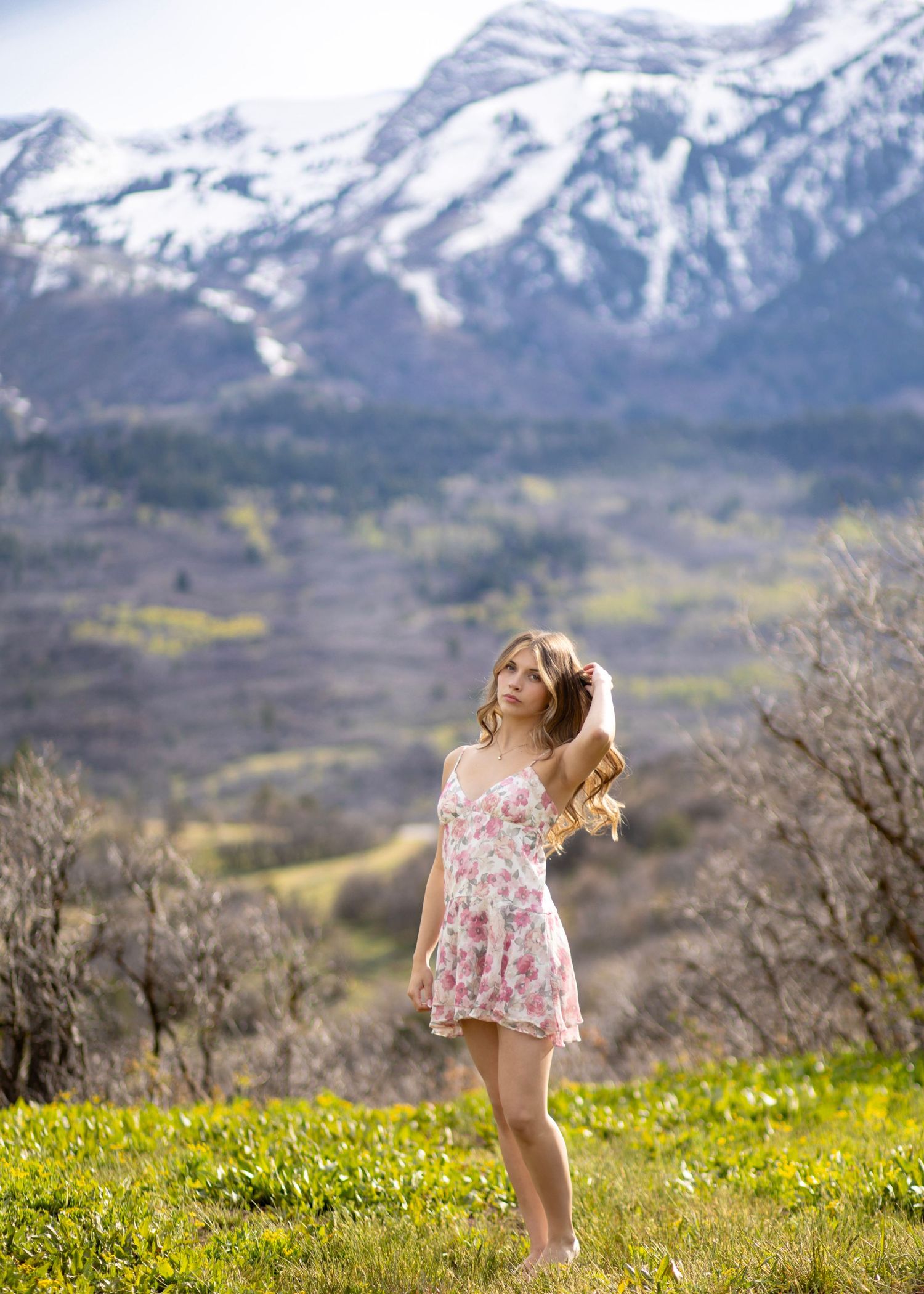 Young woman in floral dress poses on green hillside with snow-capped mountain backdrop in spring.