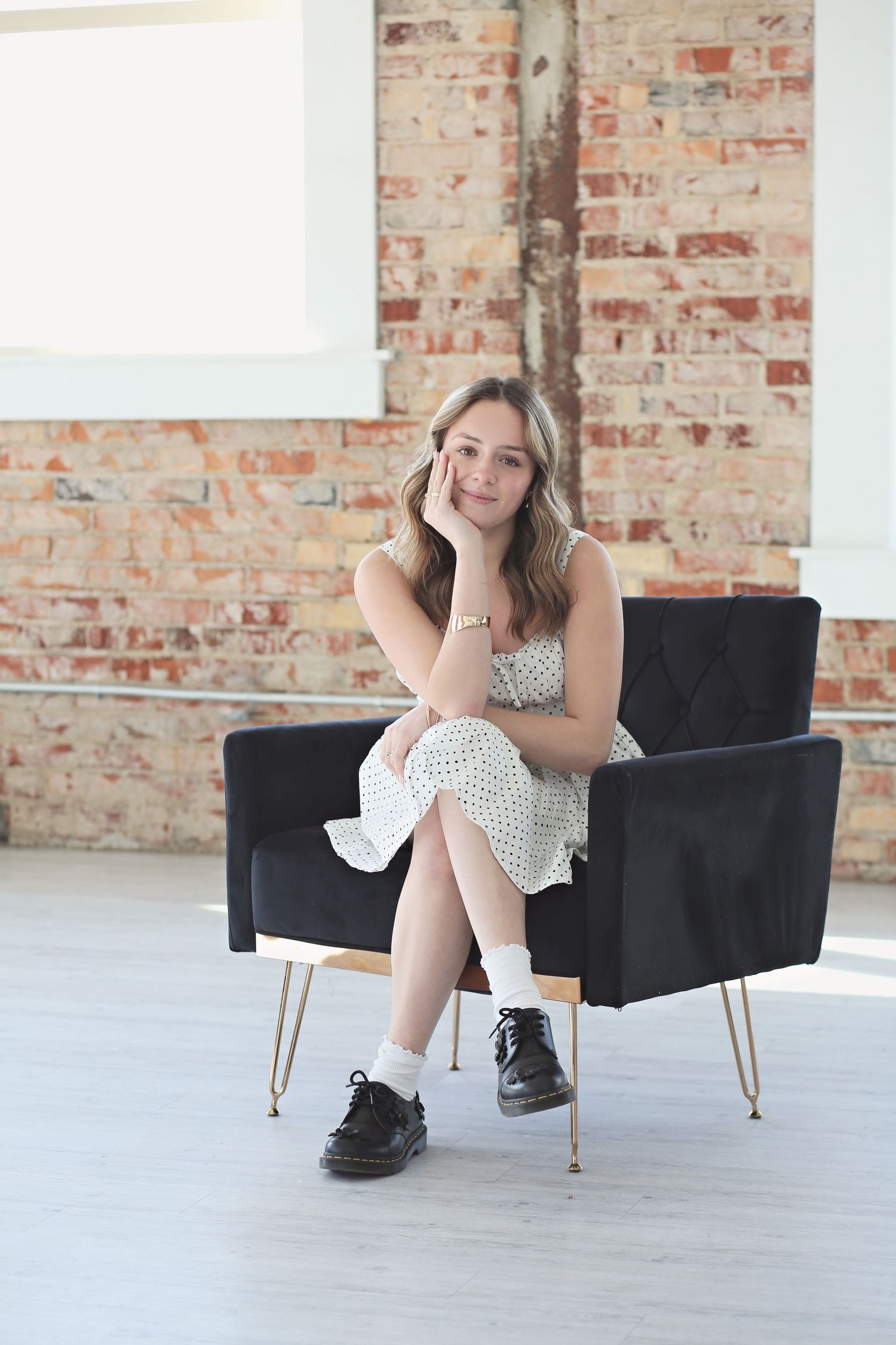 Model poses on black chair in industrial space with exposed brick walls and white floors.
