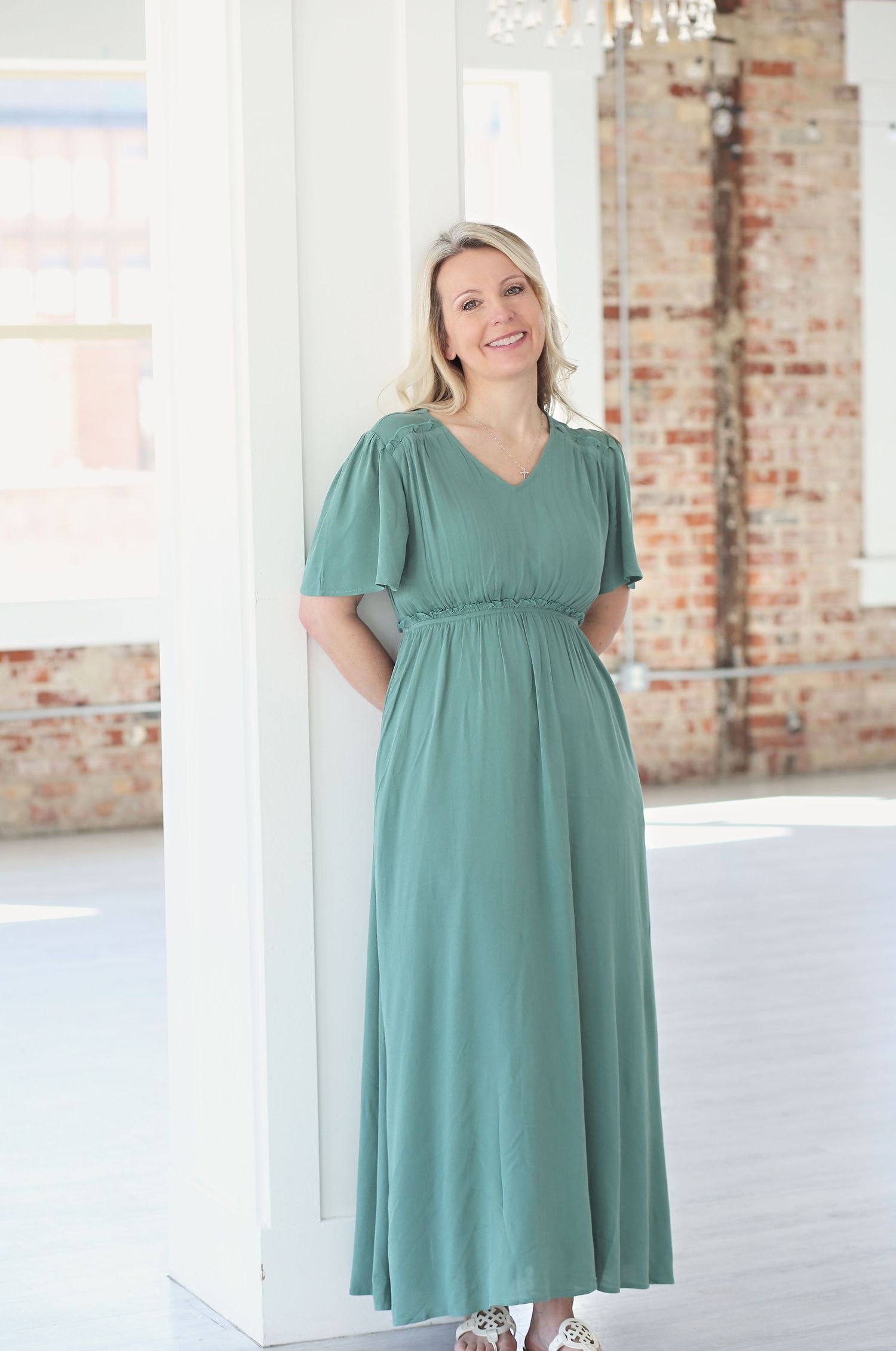 Woman wearing a flowy sage green midi dress with short sleeves and empire waist in a rustic brick studio setting.