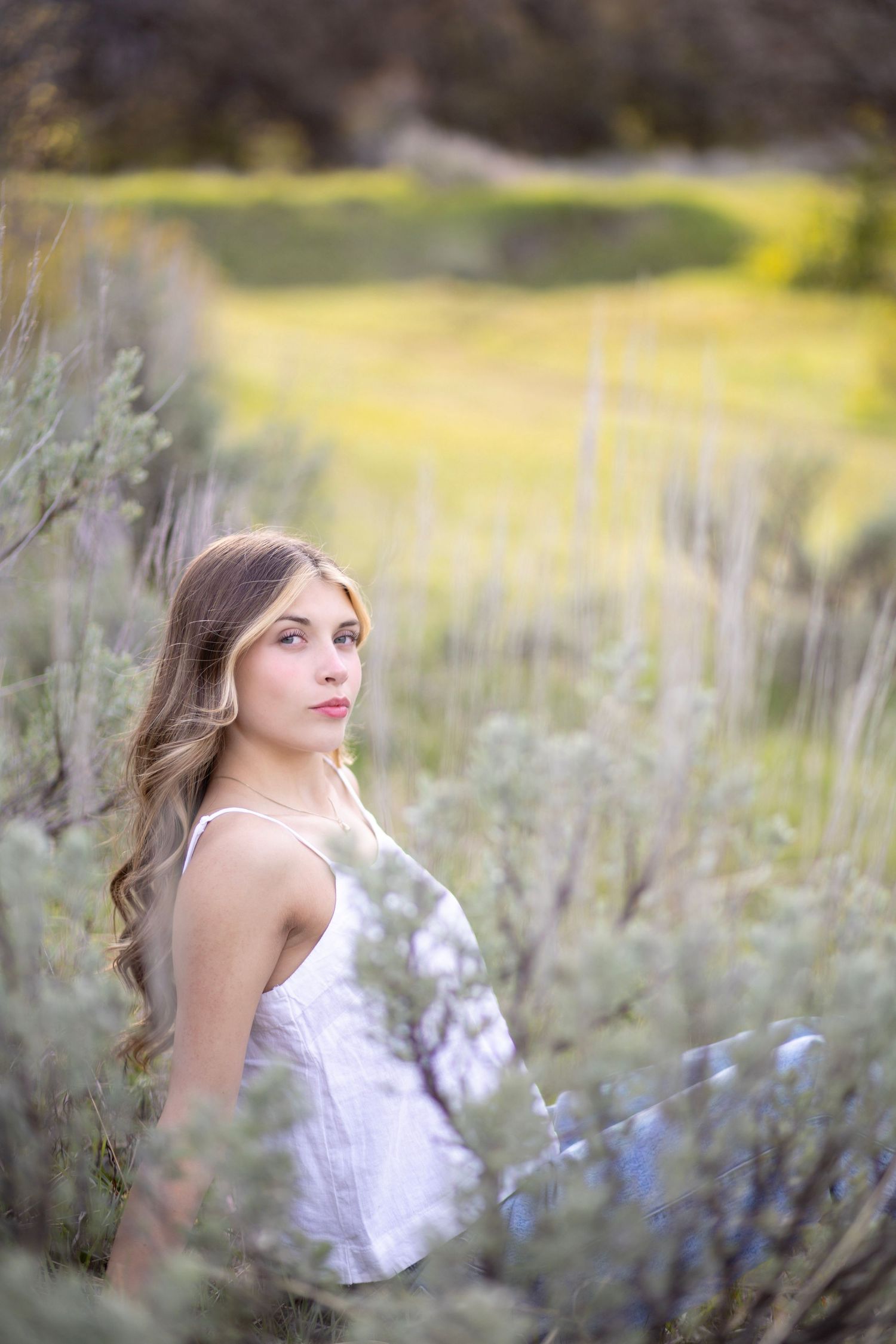 Young woman in white dress poses among sage brush in a sunlit green meadow, looking back over her shoulder.